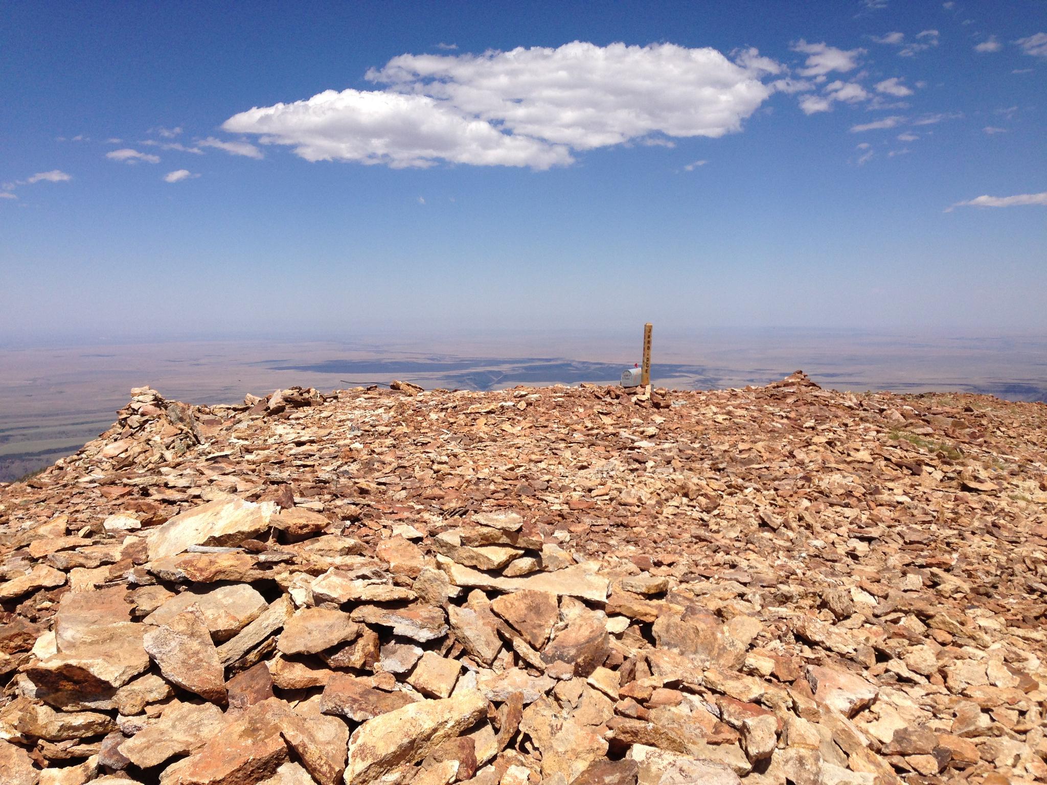 A rocky mountain summit with a wooden post under a bright blue sky.