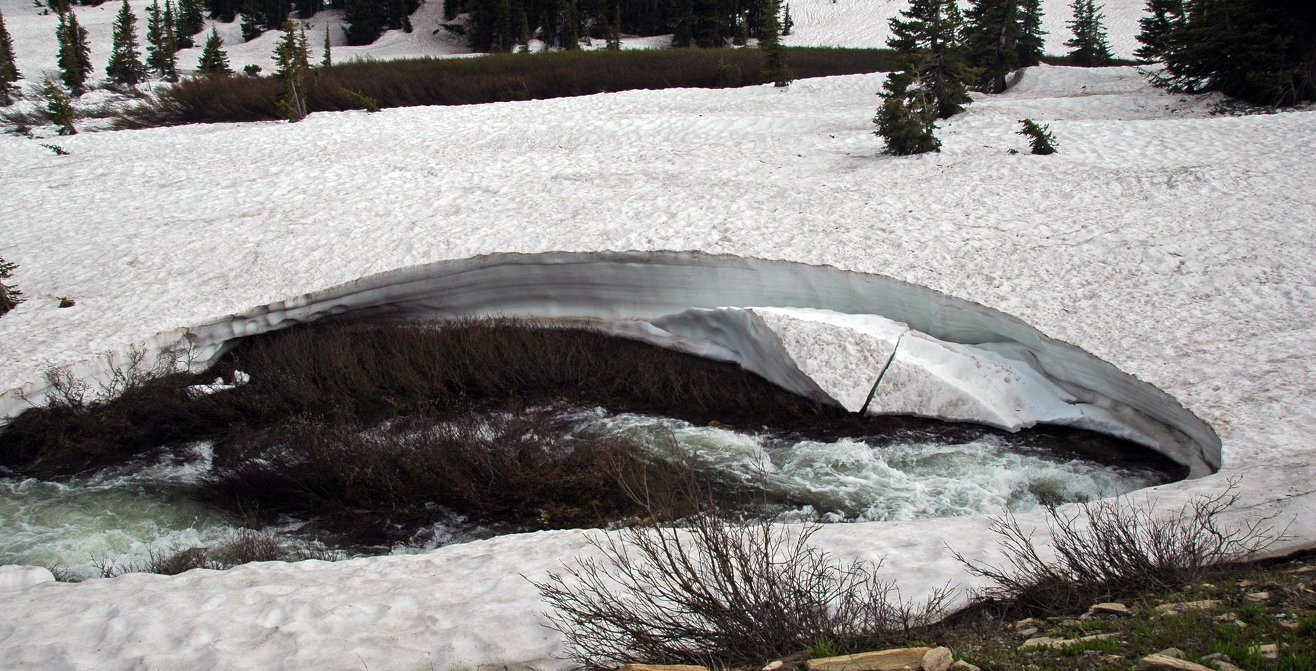A stream flows under a large snow bridge in a snowy landscape.