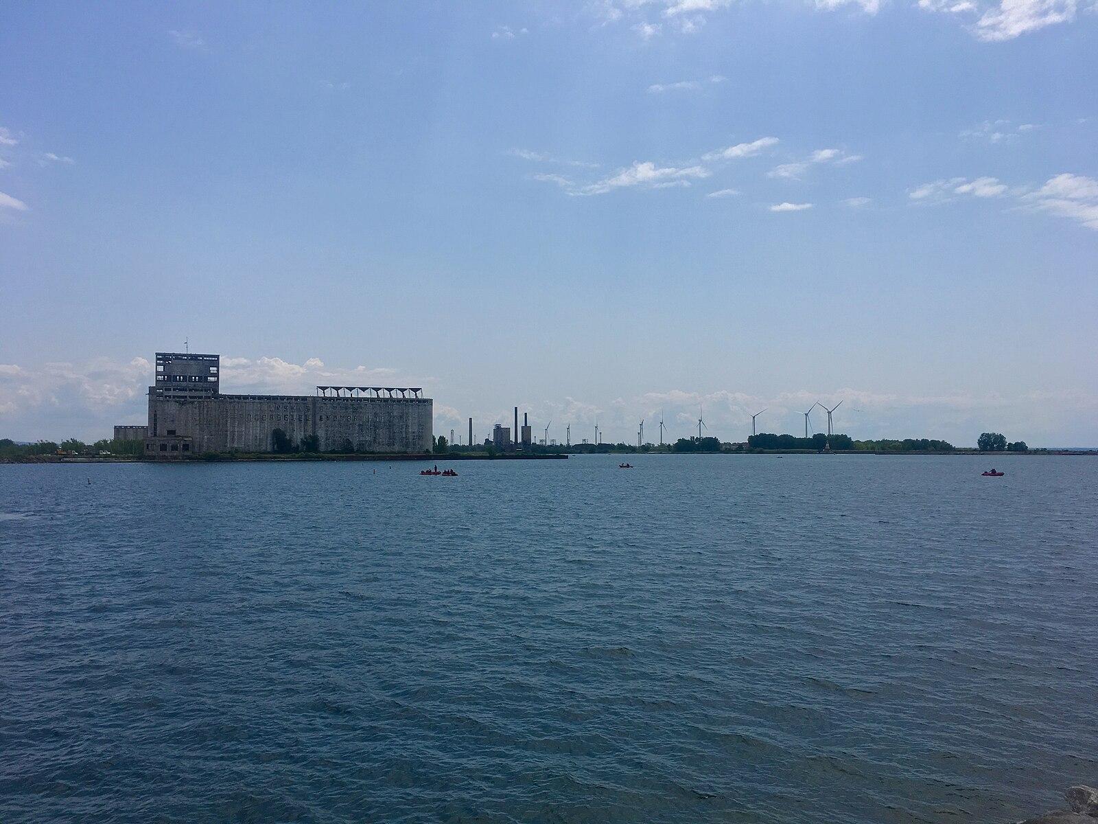 Vast body of water with a large building, wind turbines, and small boats.