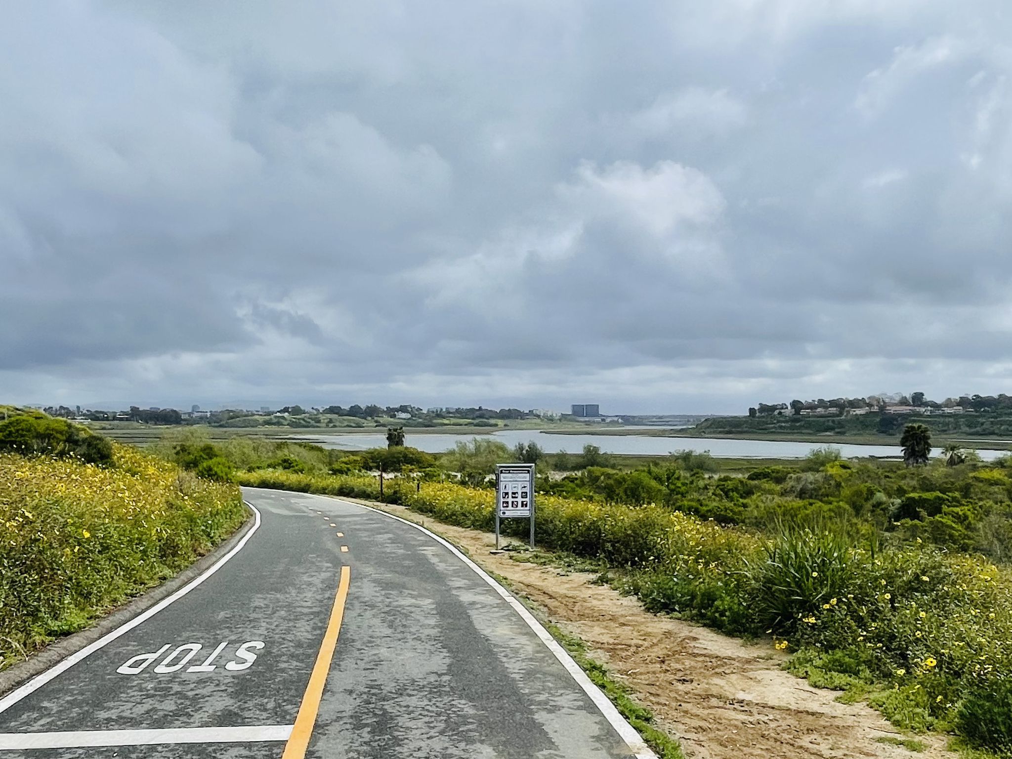 Paved path with "STOP" sign leads to a large lake under a cloudy sky. - Back Bay Loop Trail