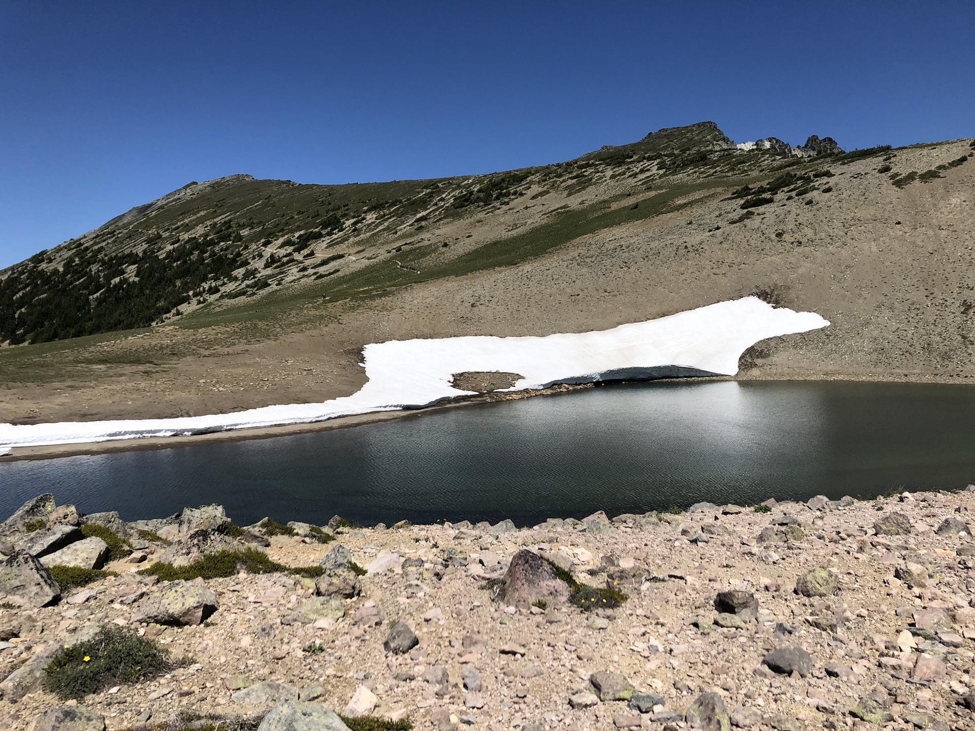 Mountain lake with a large snow patch, rocky shore, and clear sky.