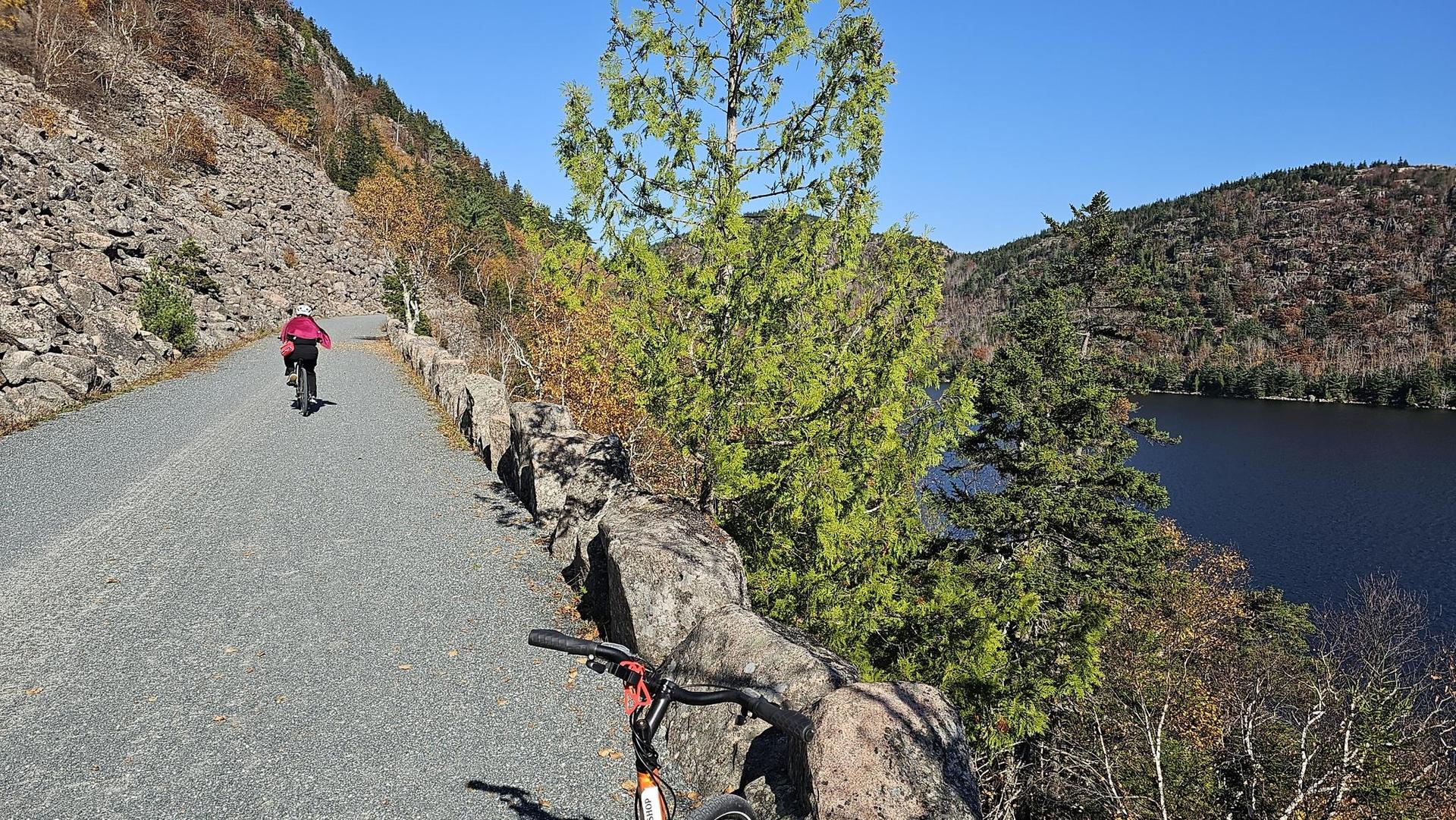Person cycling on a sunny mountain path beside a lake with autumn trees.