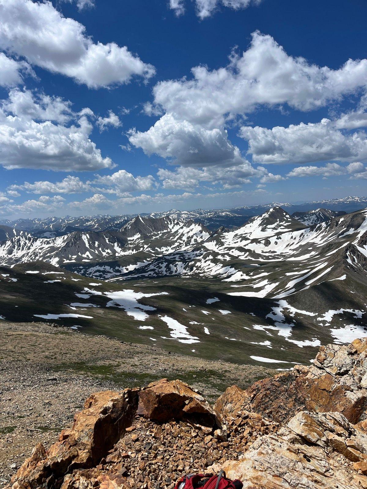 Expansive mountain vista with snow patches, green valleys, and a red backpack.