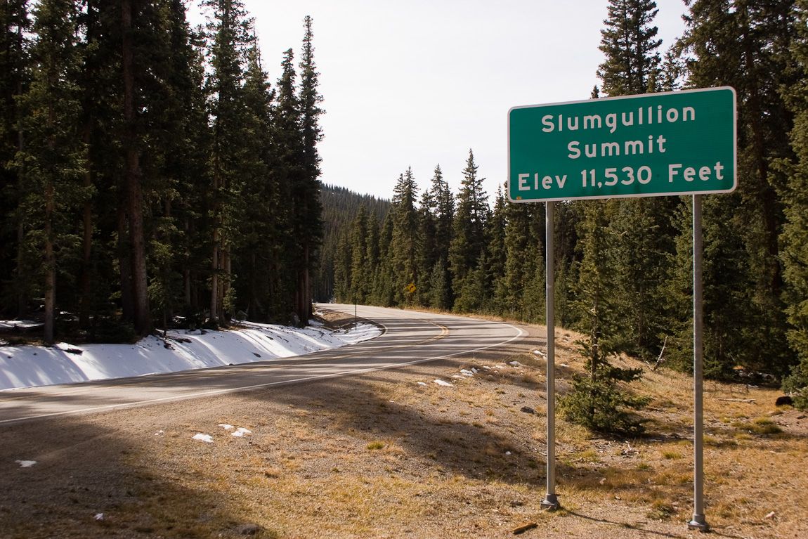 Green road sign beside a winding road through a dense pine forest with snow. - Slumgullion Pass — Lake City
