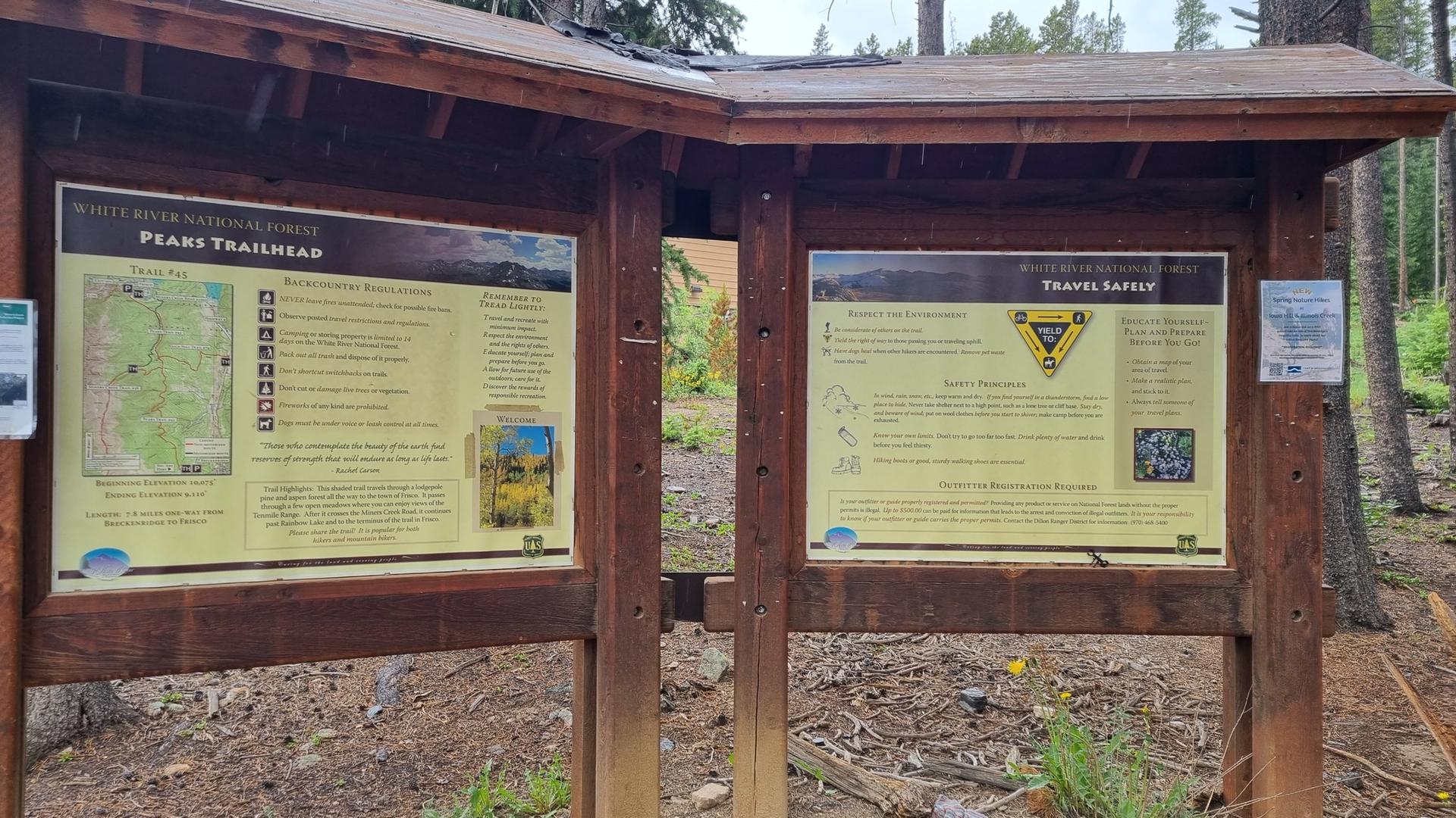 A wooden trailhead kiosk with two information signs in a forest setting.
