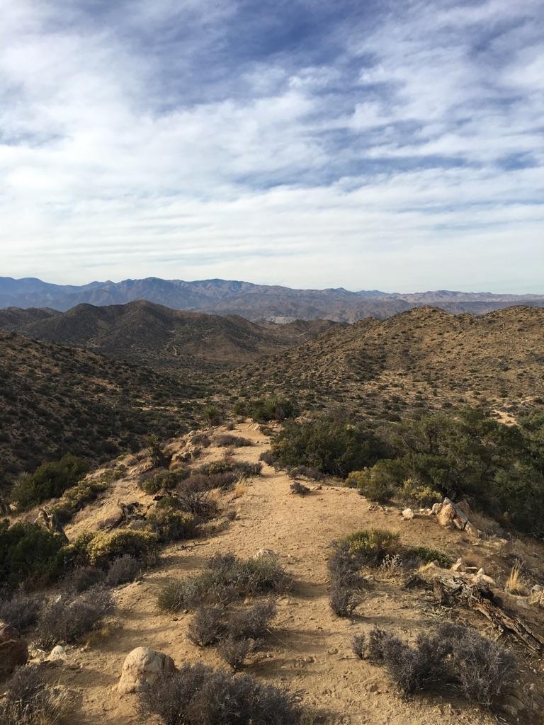 Winding dirt path on a desert ridge with rolling hills and cloudy sky.