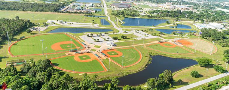 Aerial view of multiple baseball fields and ponds under bright sun.