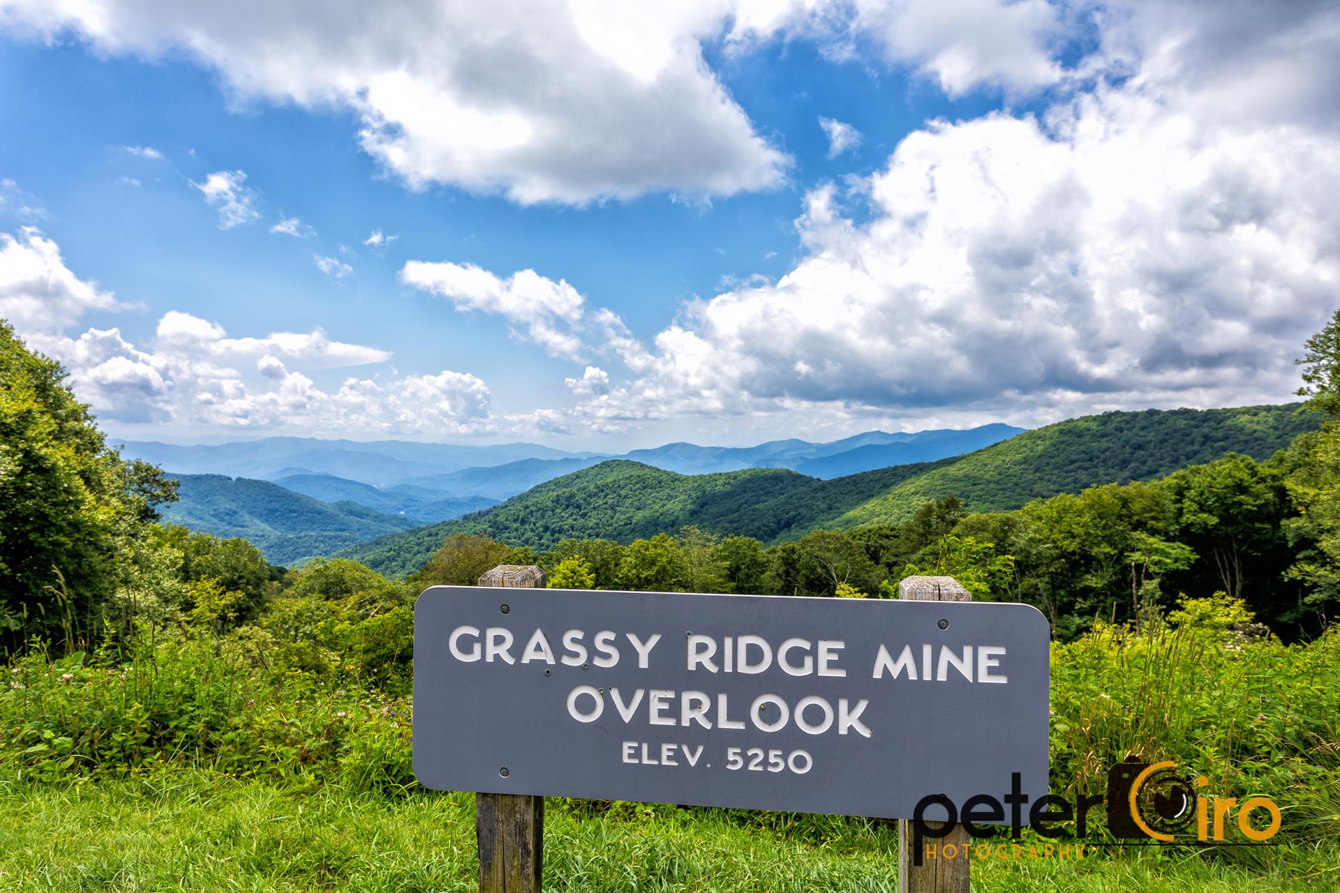 A dark sign stands in green grass overlooking lush mountains under a bright sky.