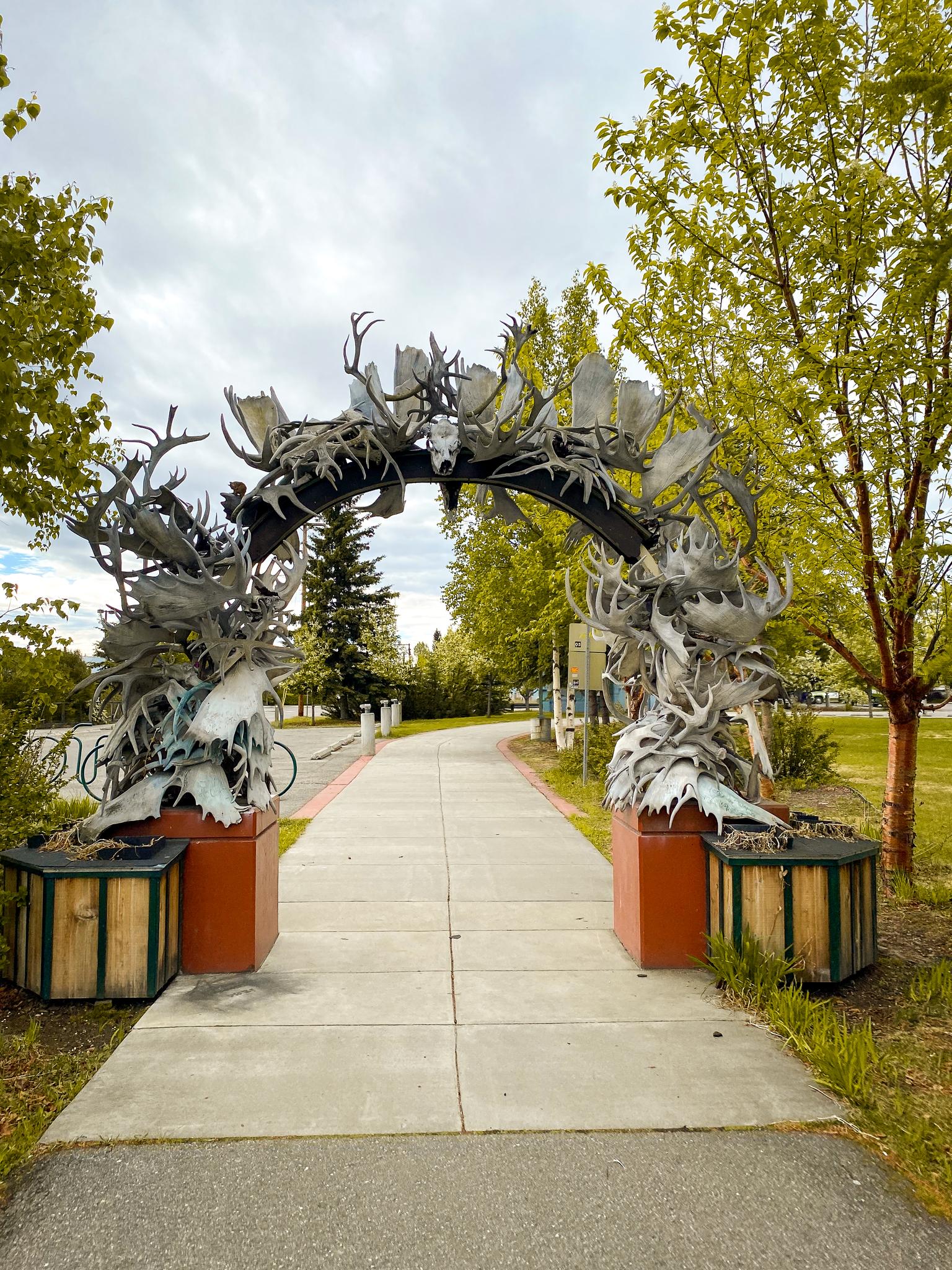 An archway made of antlers stands over a paved path, surrounded by green trees.