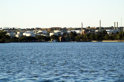 Industrial buildings and storage tanks line a distant shore across a calm body of water.