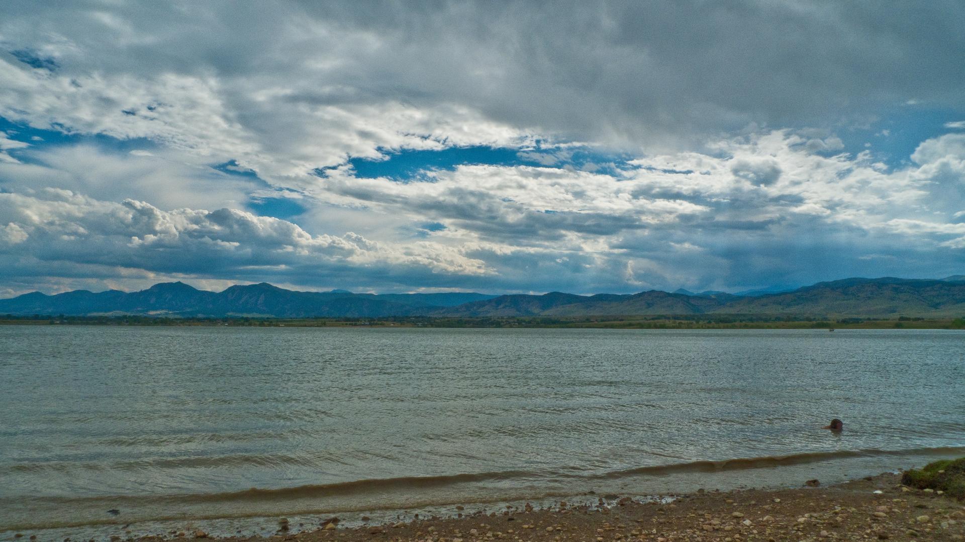 Wide lake under cloudy sky with distant mountains and a rocky shore.