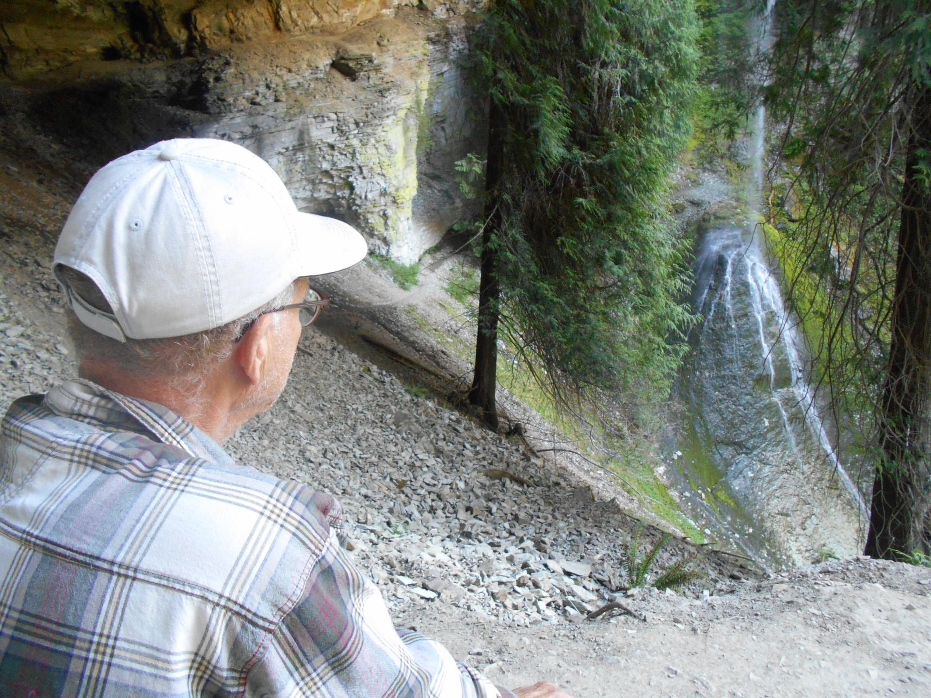 Back of a person in a cap observing a waterfall in a shaded grotto.