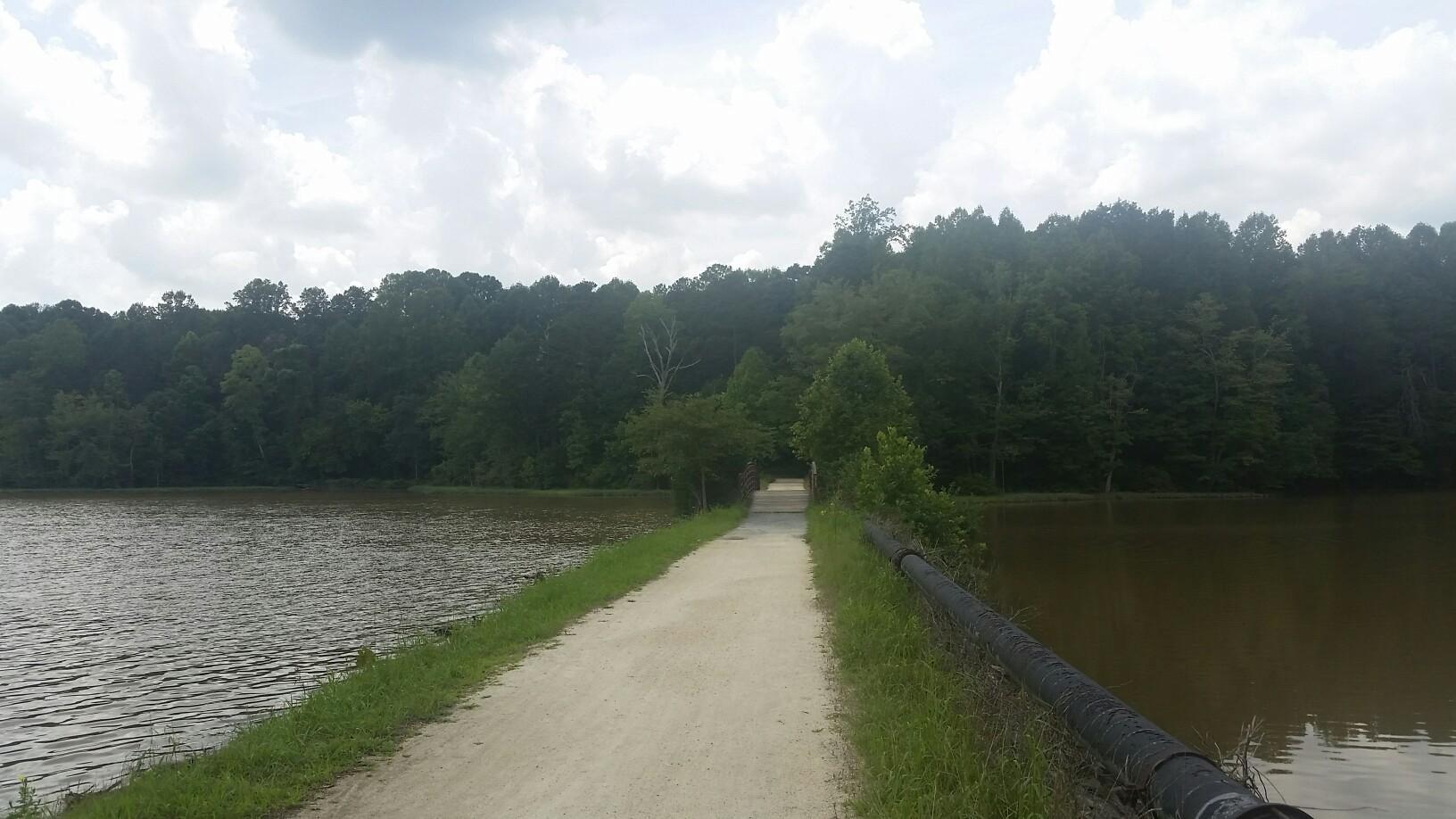 Overcast day, a dirt path divides two lakes with a dark forest beyond.