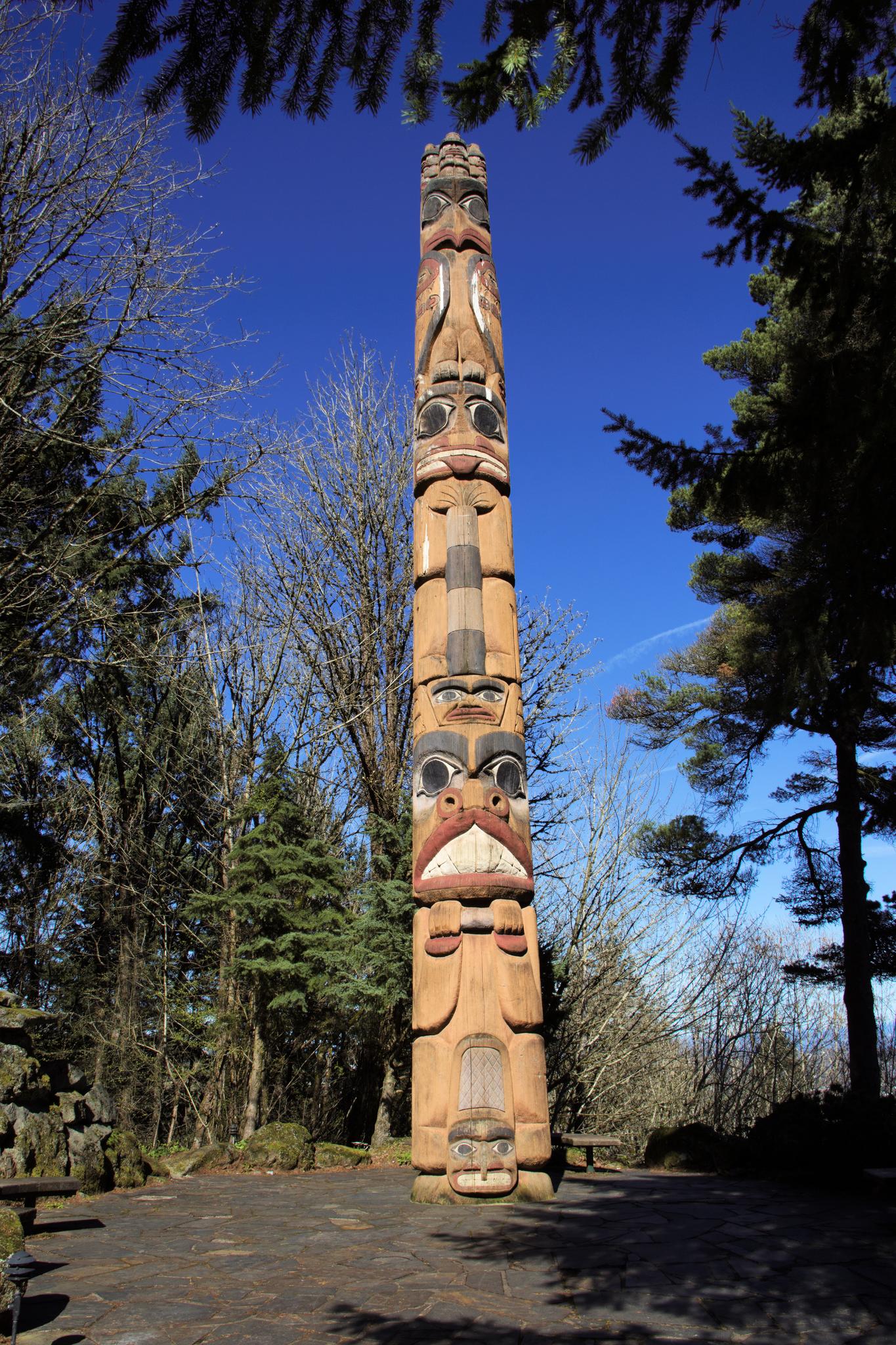 Tall, intricately carved totem pole stands under a bright blue sky among trees.