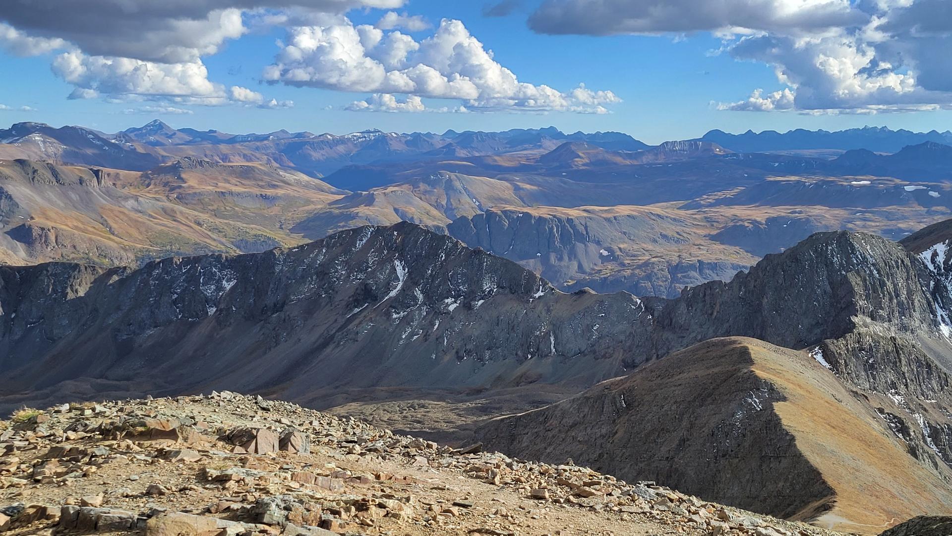 Expansive mountain landscape with rocky peaks, valleys, and scattered snow under a cloudy sky.