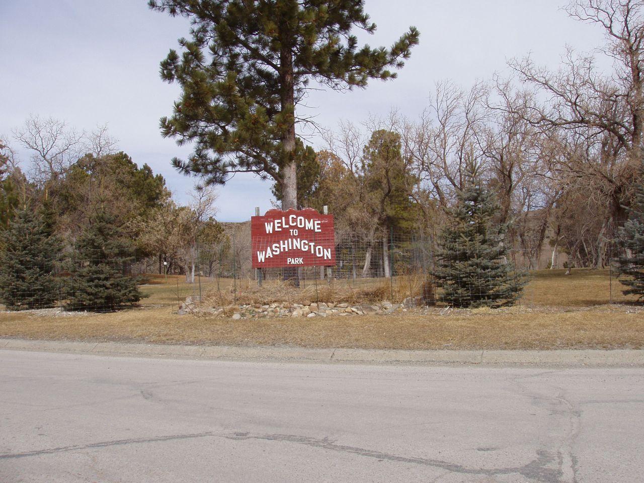 A red welcome sign stands among trees and grass beside a paved road.