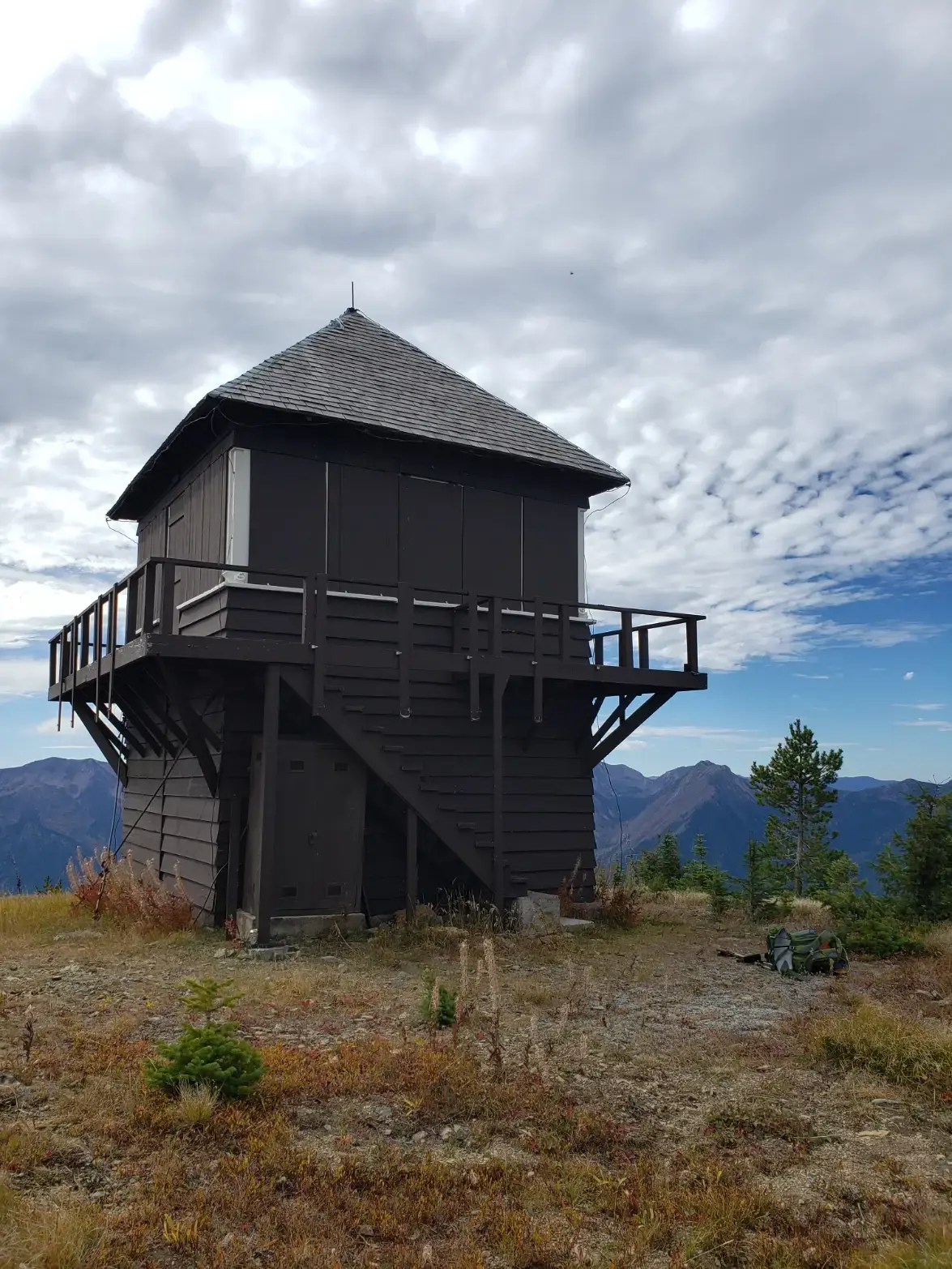 Dark wooden fire lookout cabin on a mountain ridge under cloudy skies.
