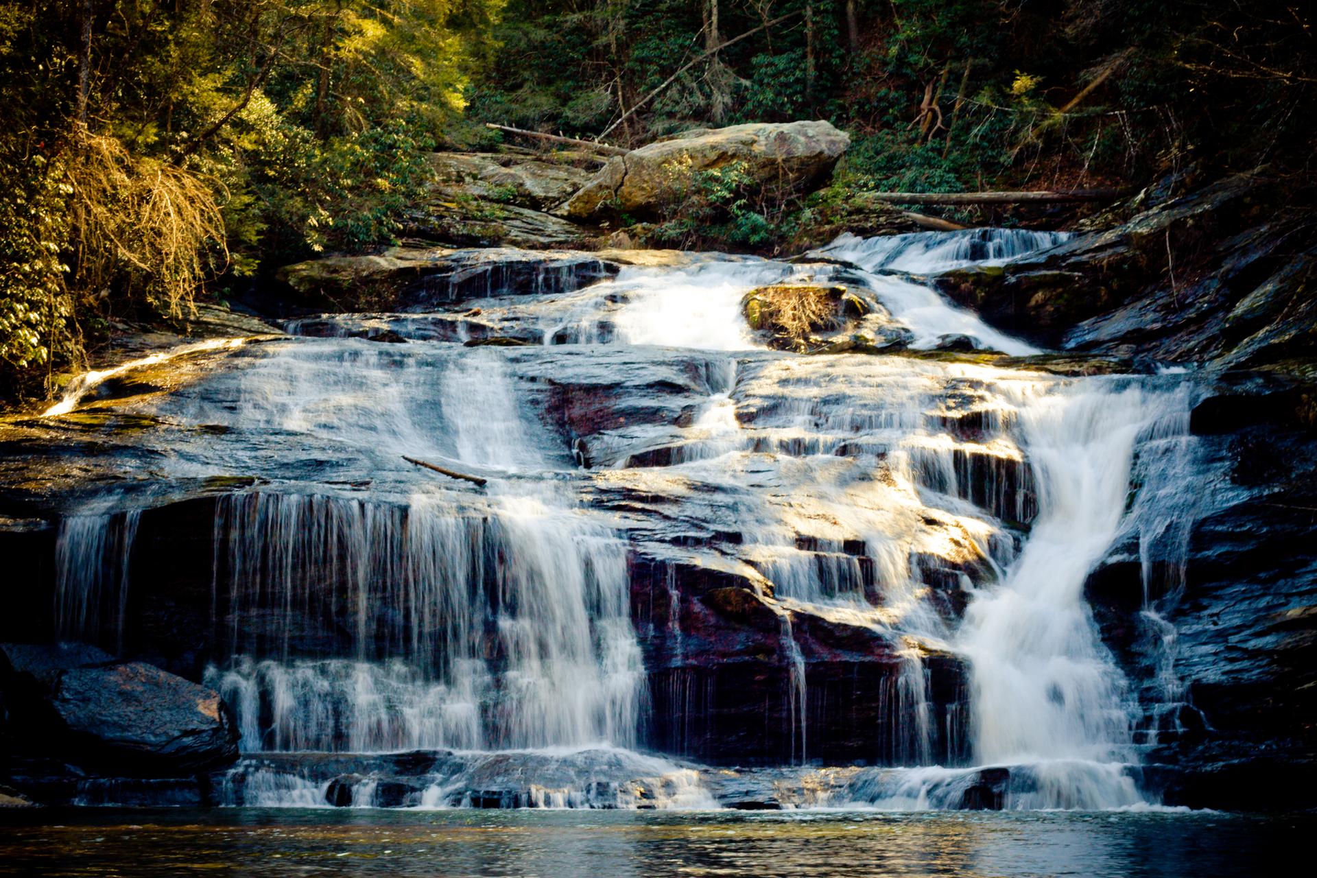 Sunlight illuminates a beautiful cascading waterfall surrounded by green trees and rocks.