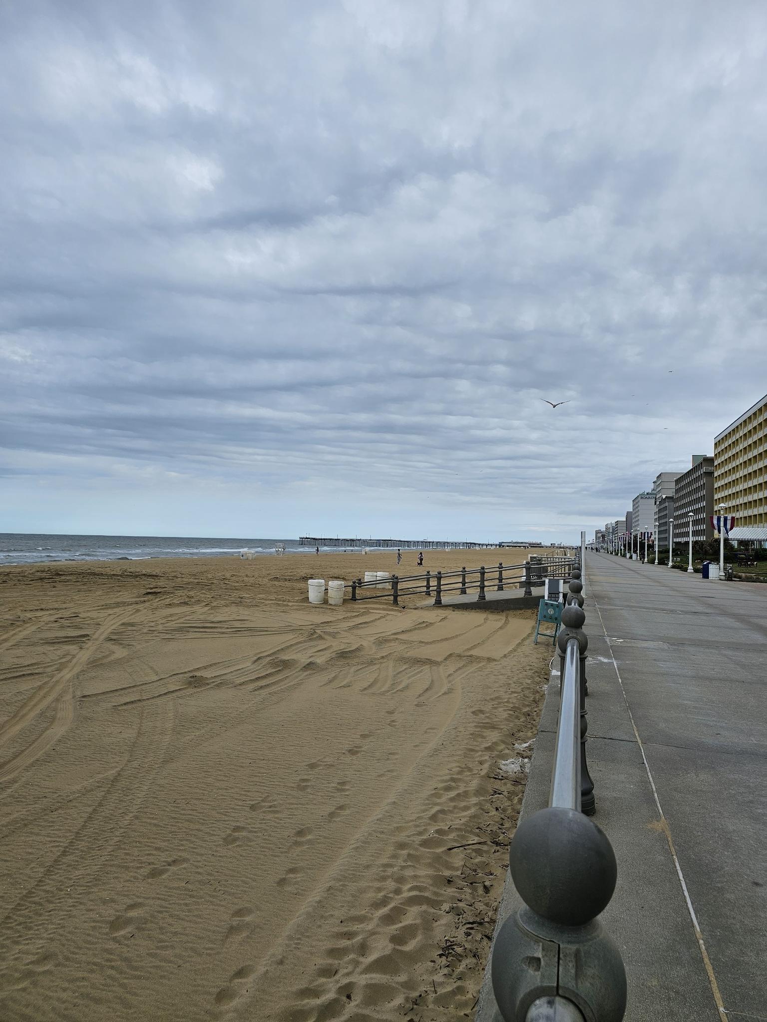 A wide sandy beach and boardwalk under a cloudy sky, with distant buildings.