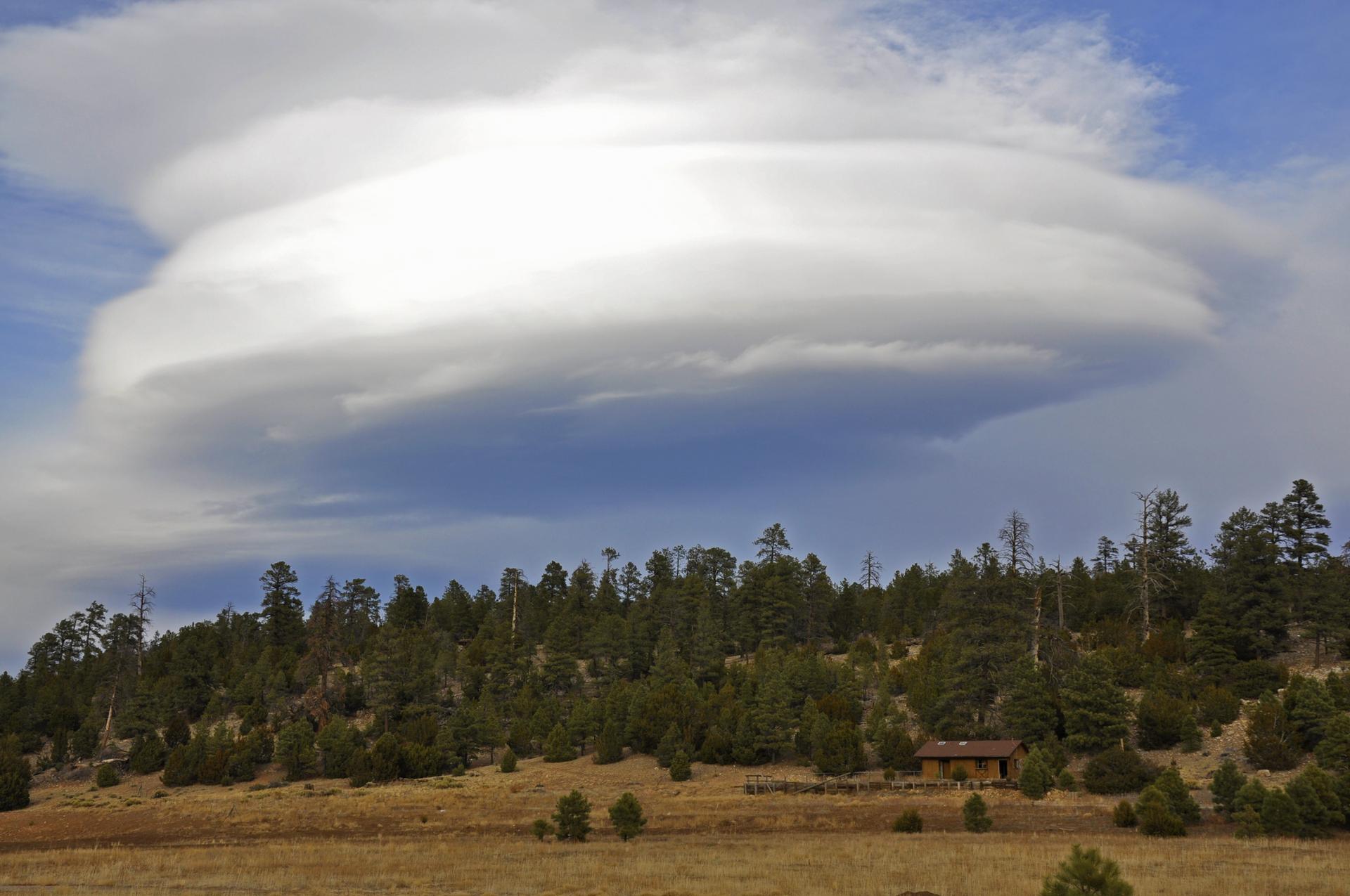 Dramatic lenticular cloud above a forest and dry field with a small cabin.