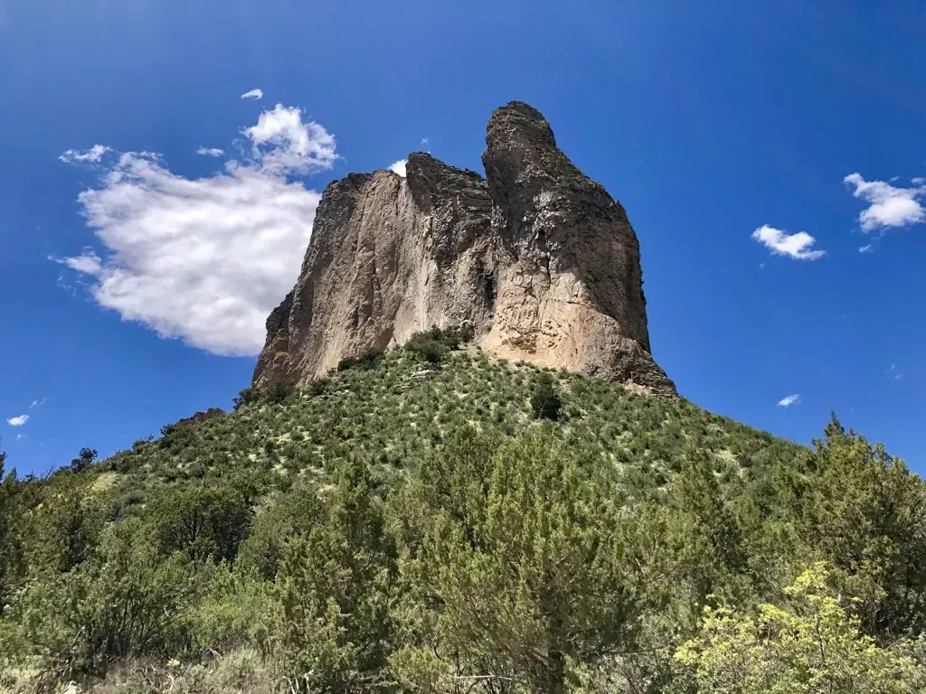 Towering rock formation rises above green scrubland under a bright blue sky. - Needle Rock Trail
