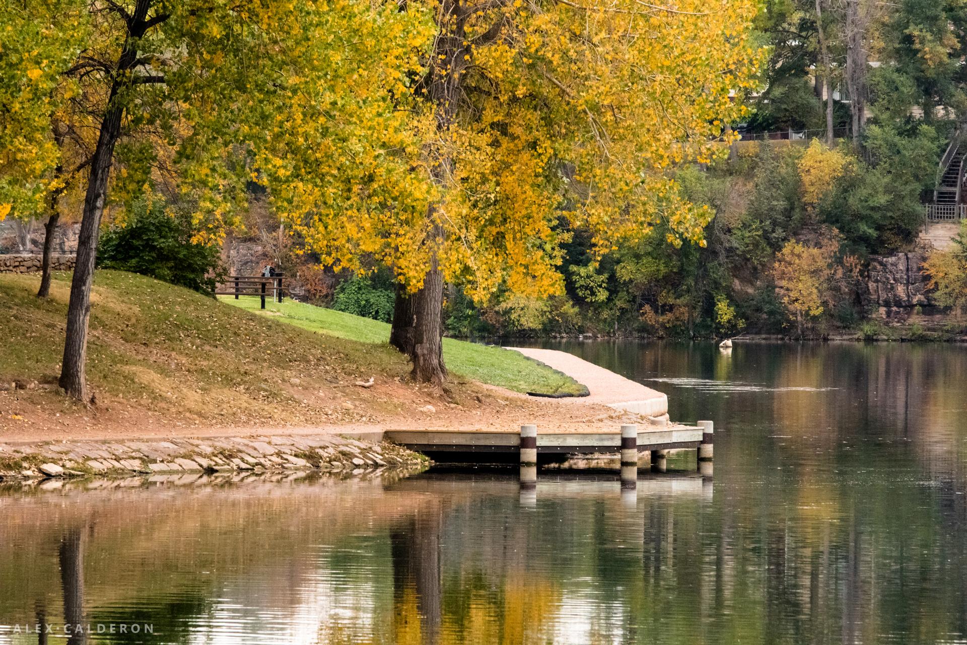 Wooden dock on a calm lake, surrounded by trees with yellow autumn leaves.