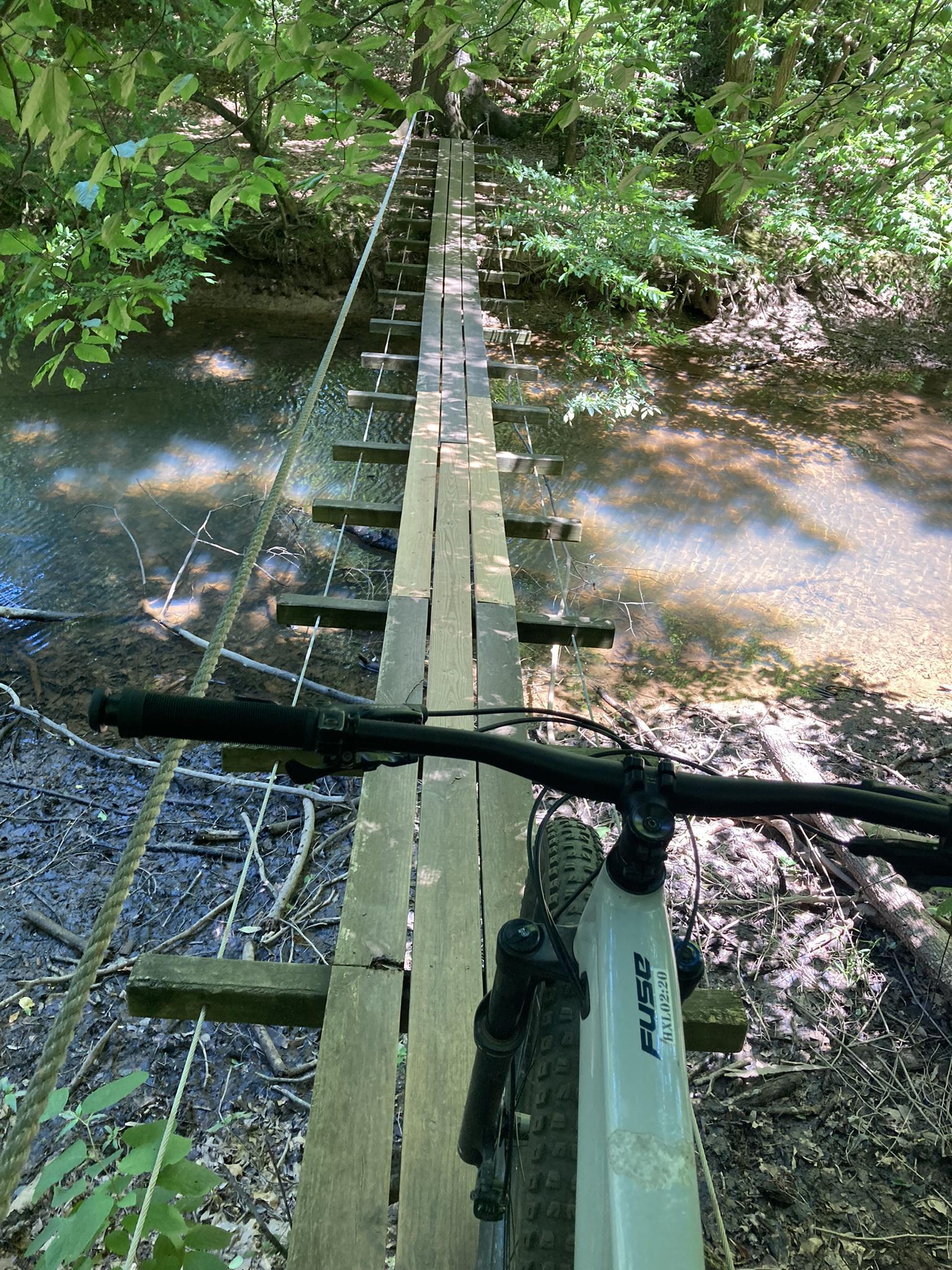 Mountain bike on a narrow wooden suspension bridge over a stream in a sunny forest.