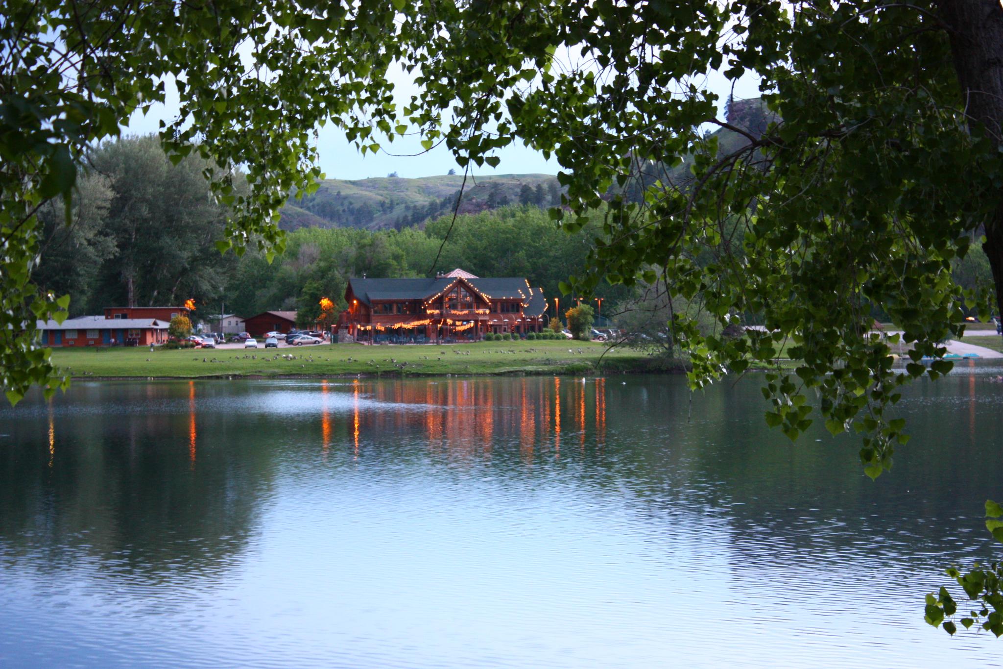 Buildings with lights reflecting on a tranquil lake at twilight, framed by leaves.