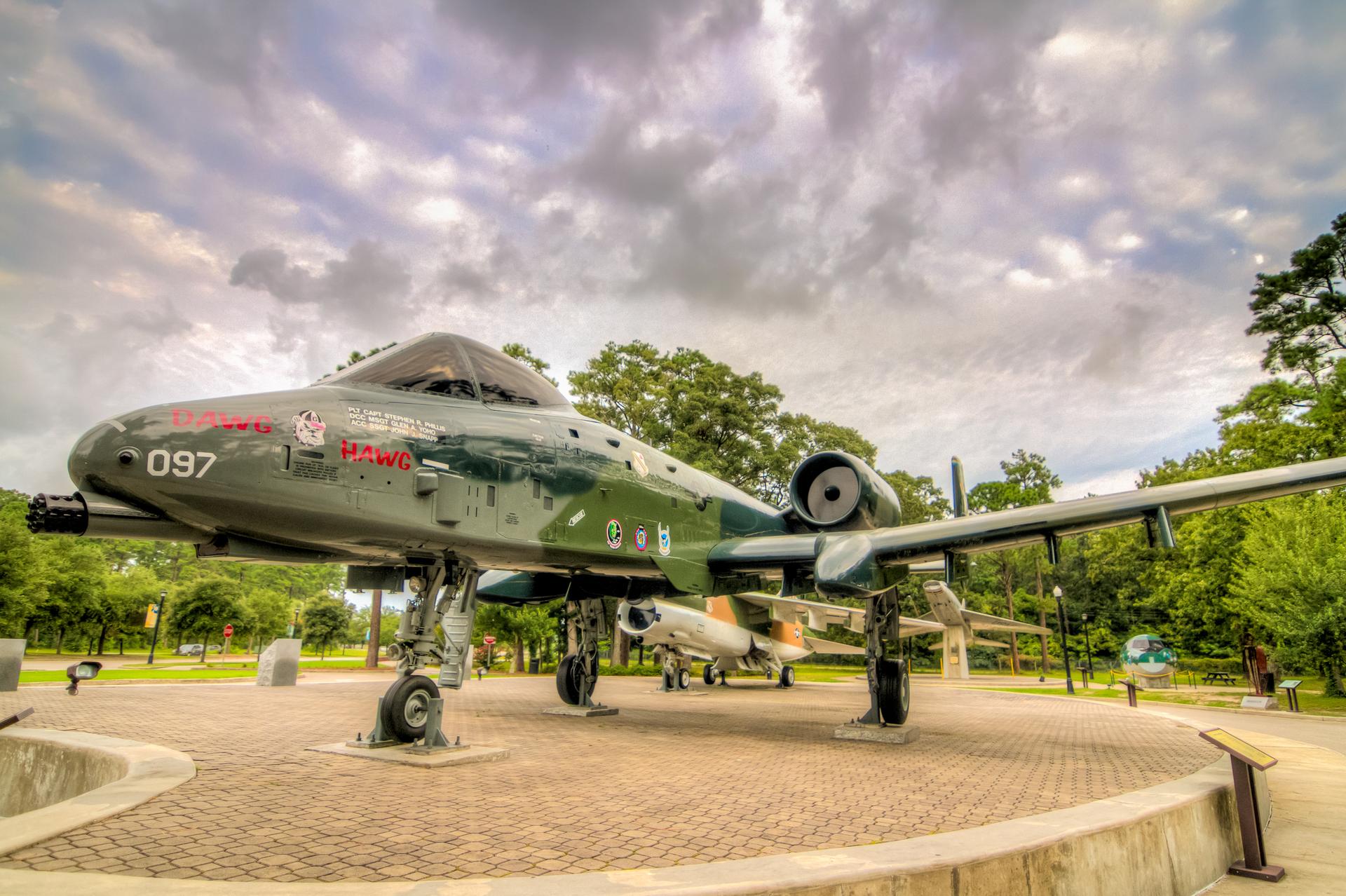 A green military aircraft on display outdoors under a cloudy sky.