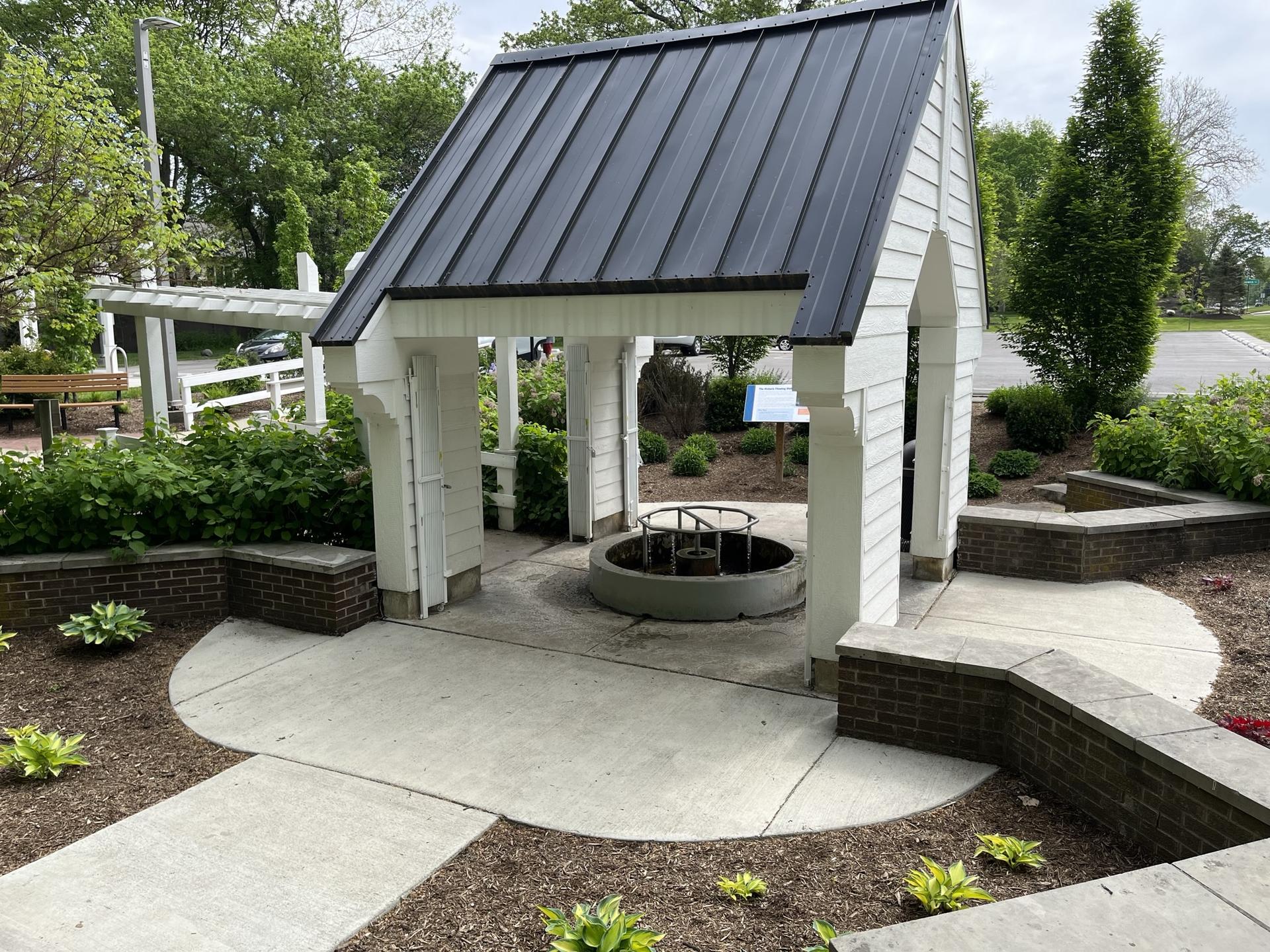 White gazebo with dark roof, circular well, and green garden beds.
