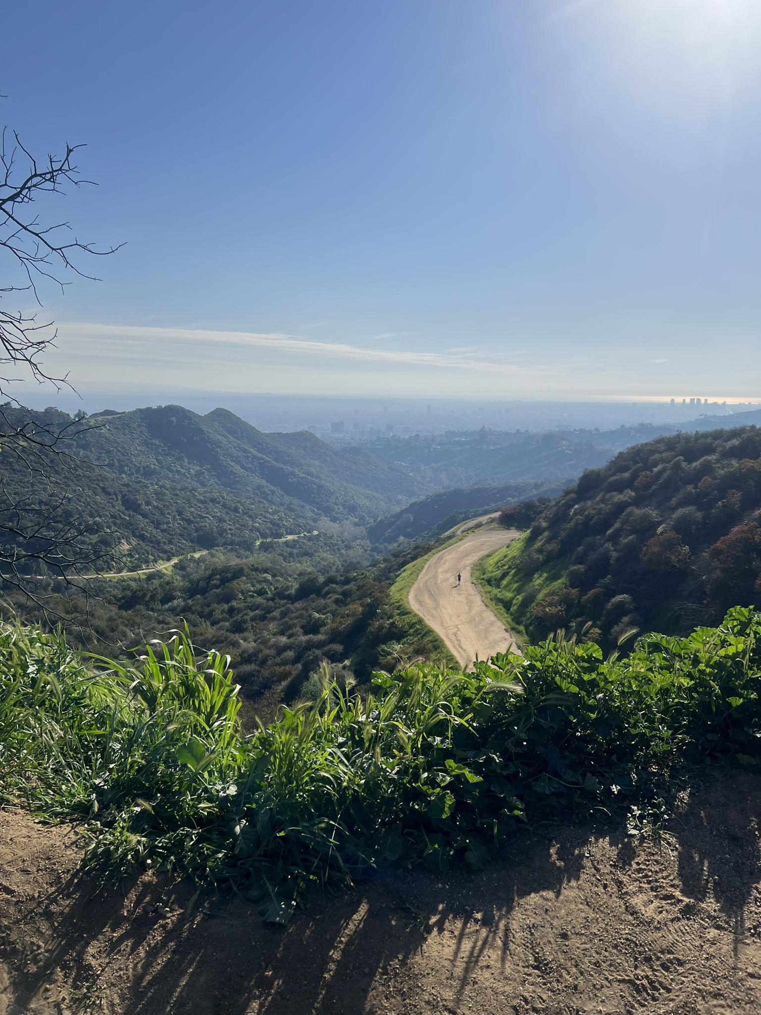 A sunny overlook shows a green valley, winding dirt road, and distant city.