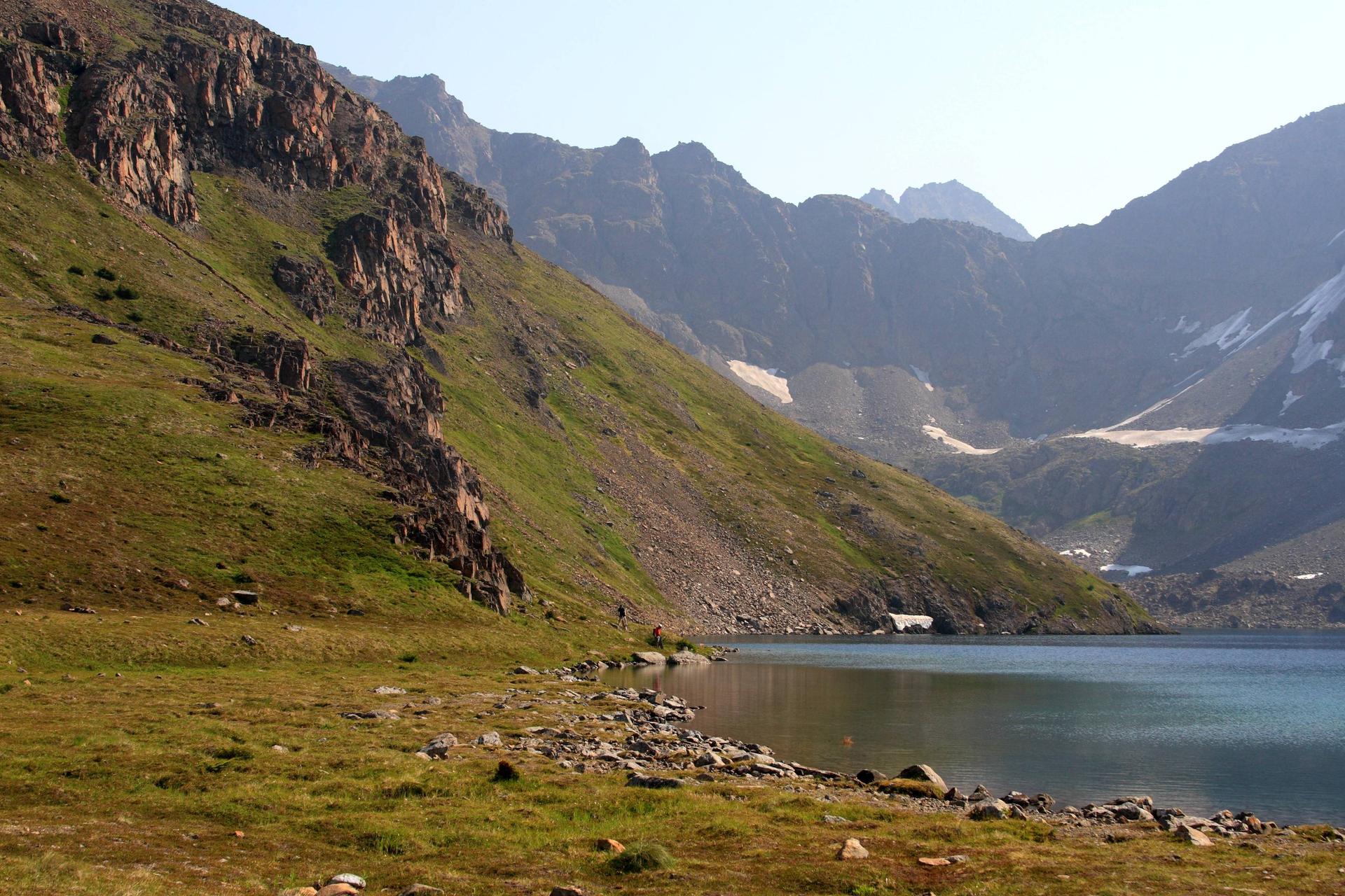 A clear lake at the base of steep, rocky, green mountains in bright light.