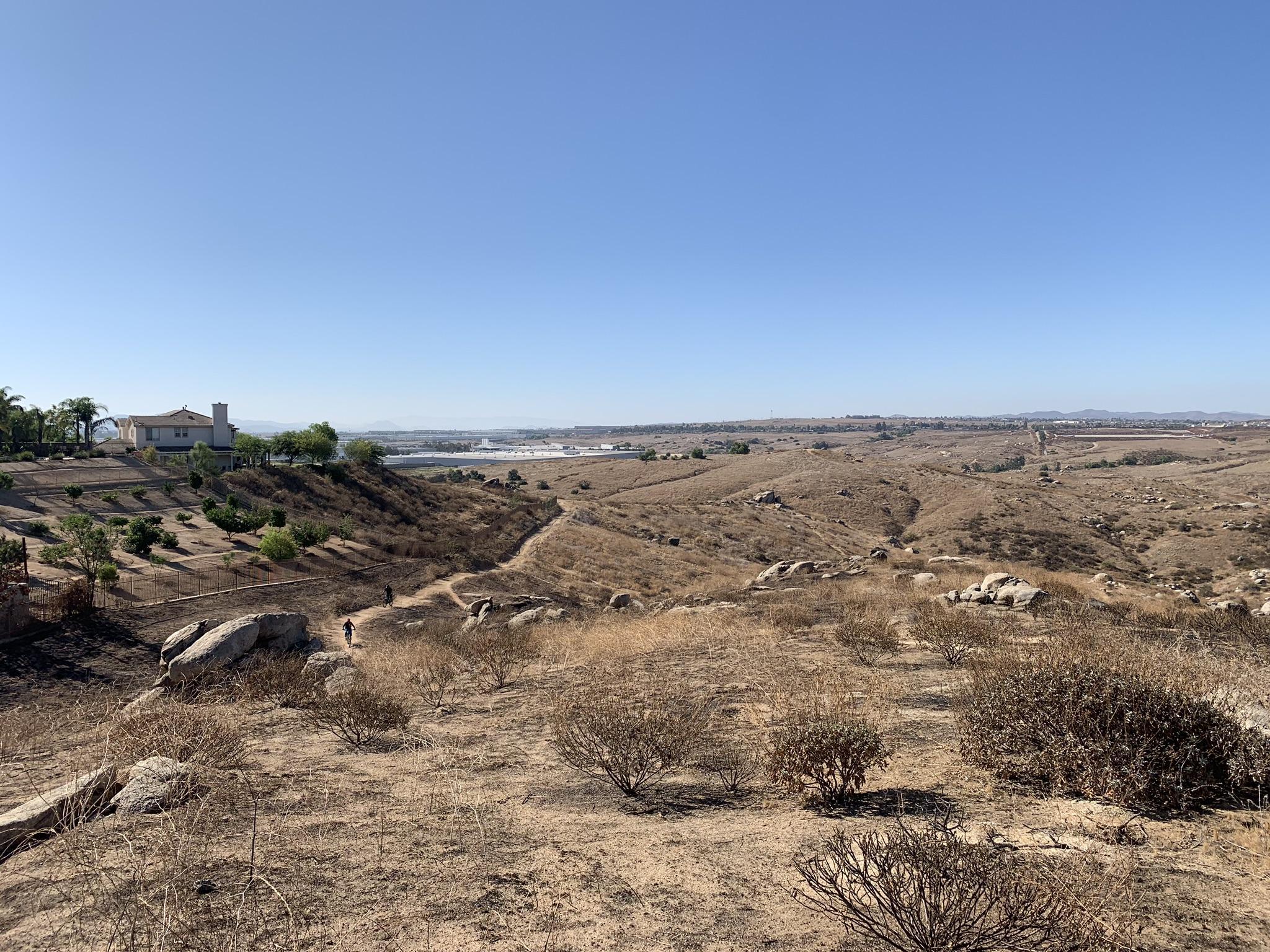 Expansive dry hills under a clear sky with distant buildings and hikers.