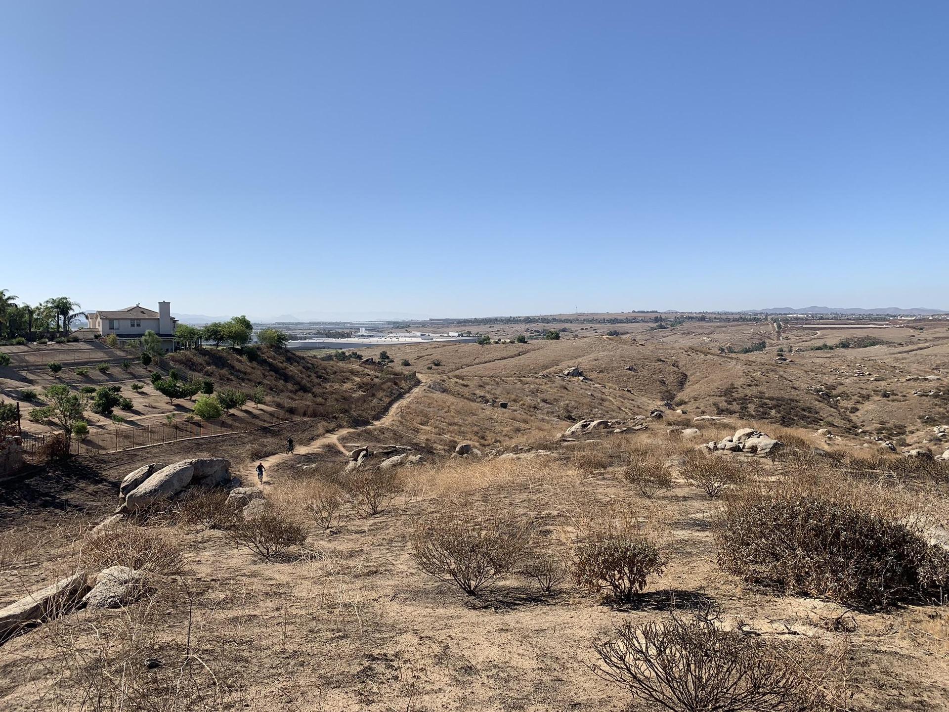 Expansive dry hills under a clear sky with distant buildings and hikers.