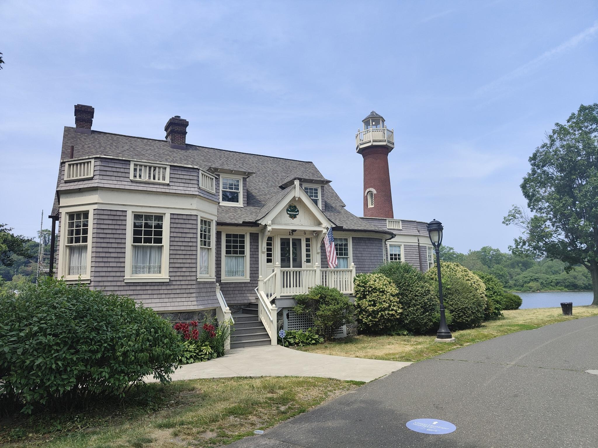 Large house with lighthouse tower, green bushes, water in background, bright day.