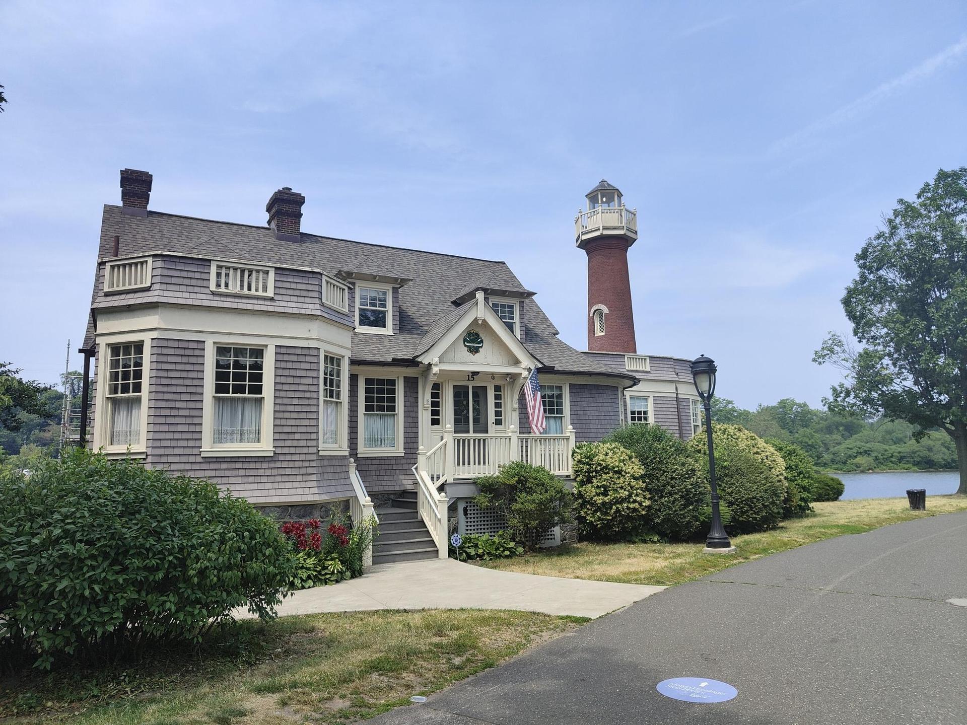 Large house with lighthouse tower, green bushes, water in background, bright day.