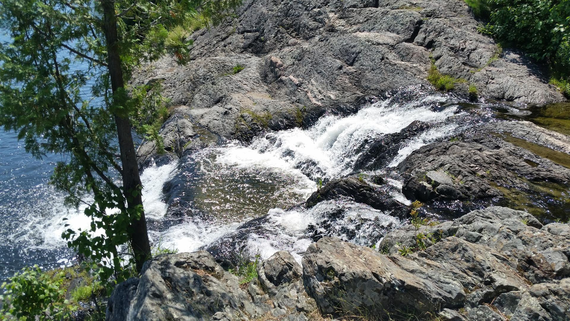 Bright waterfall cascading over dark rocks into a blue river, with green foliage.