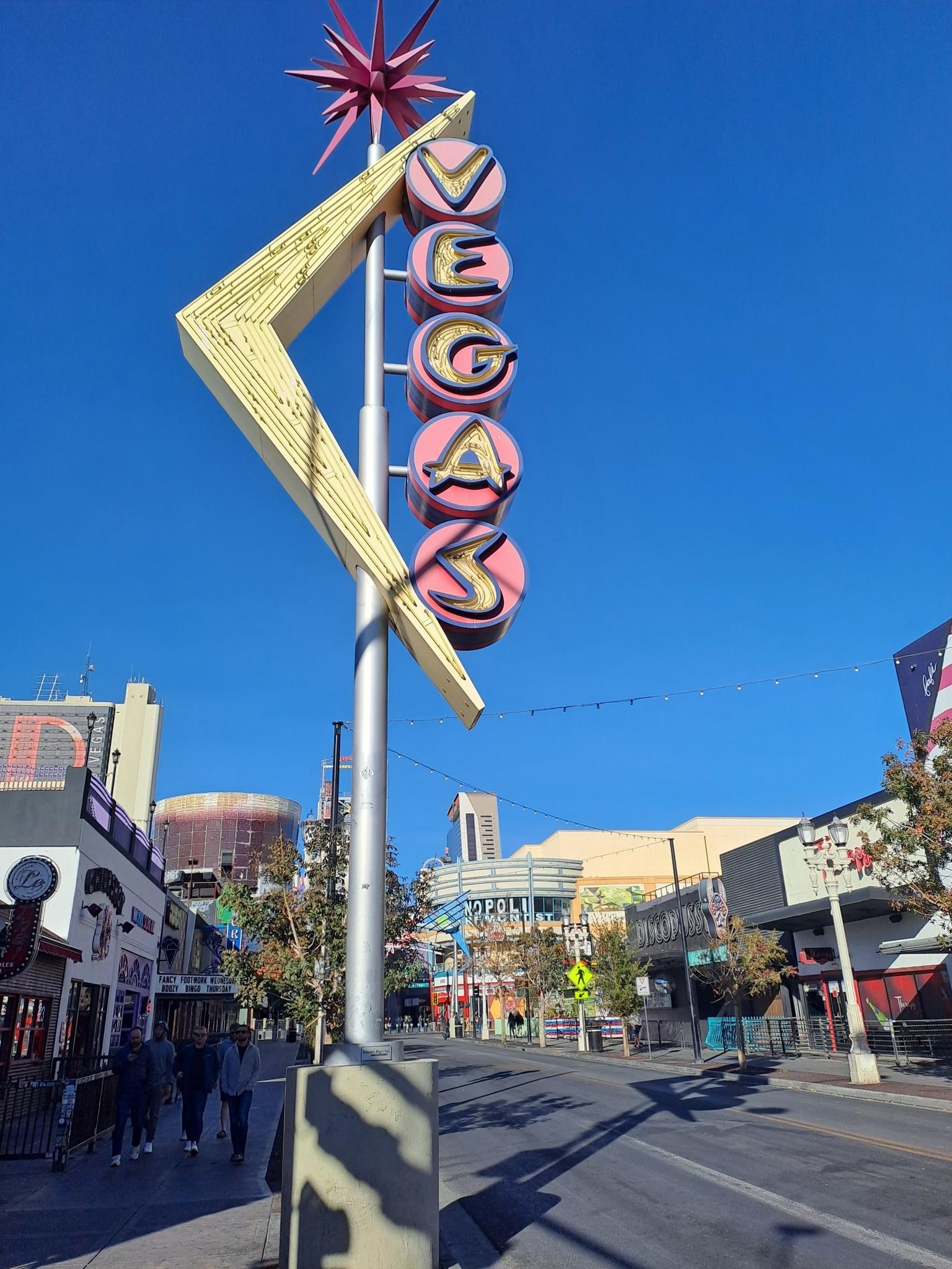 Retro 'VEGAS' sign stands tall on a sunny urban street with pedestrians.