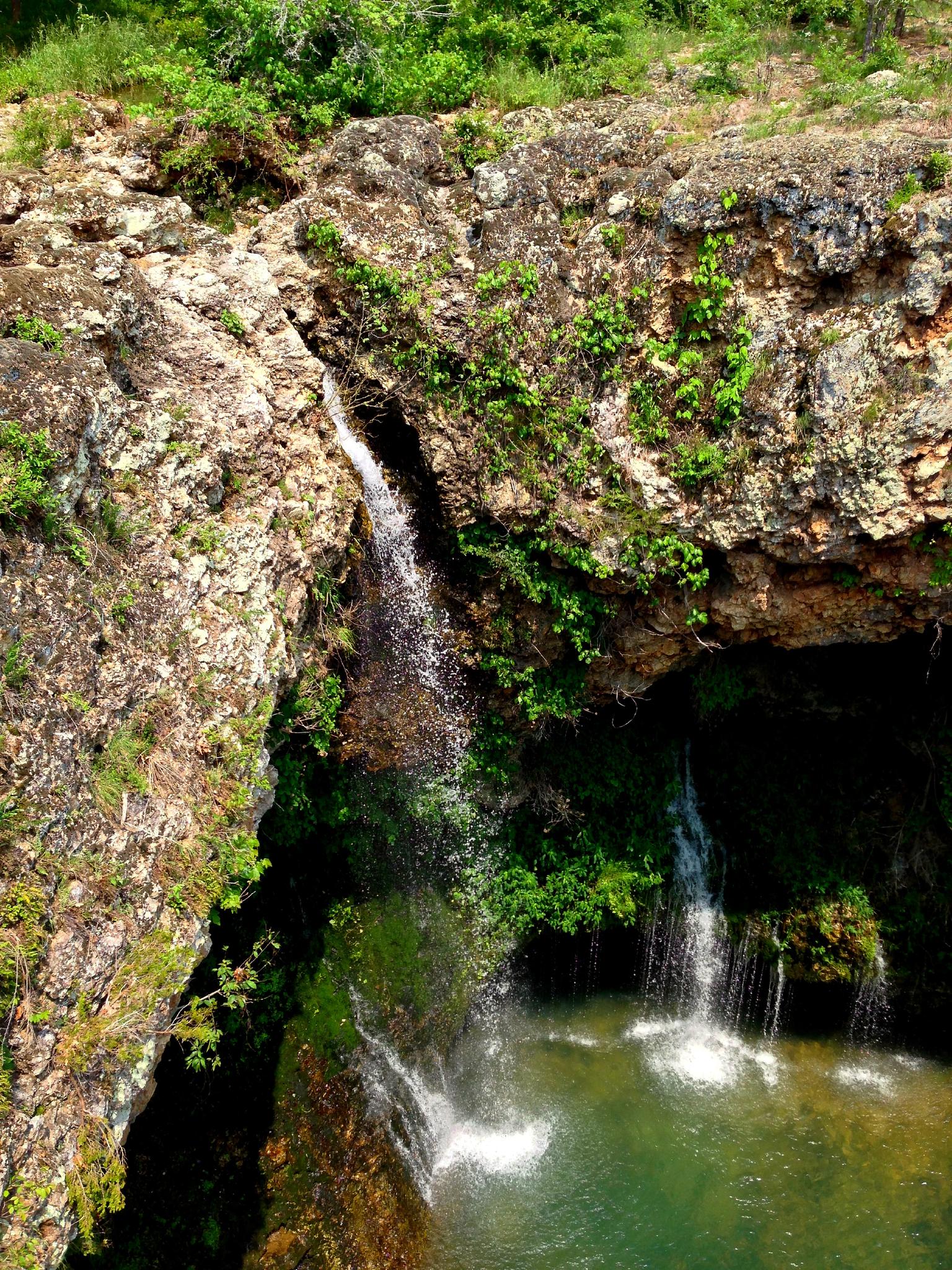 Multiple waterfalls cascade into a green pool surrounded by rocky, mossy cliffs.