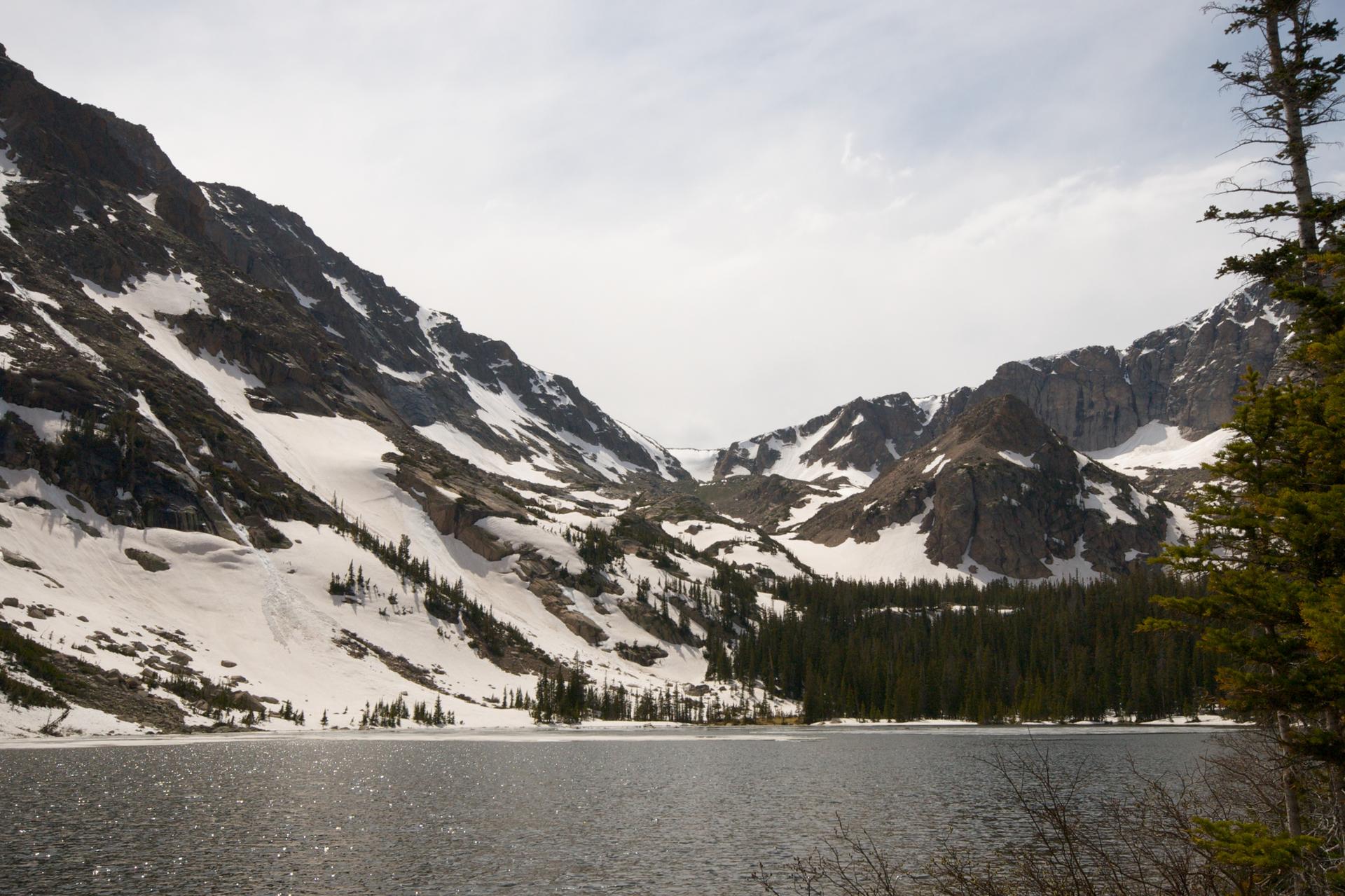 Alpine lake surrounded by snow-covered mountains and evergreen trees under cloudy sky.