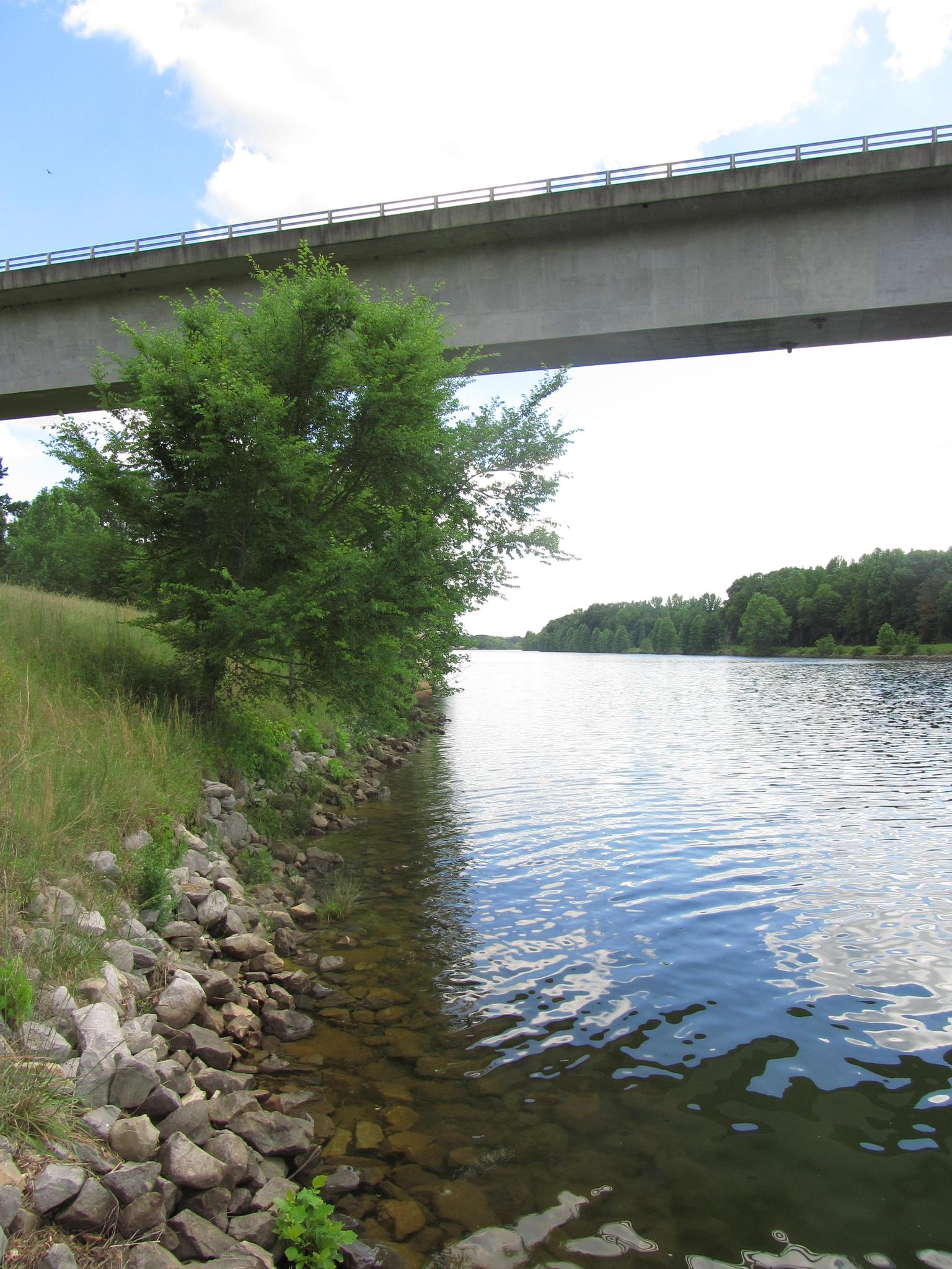 Concrete bridge spanning over water with a rocky shore and green trees on a bright day.