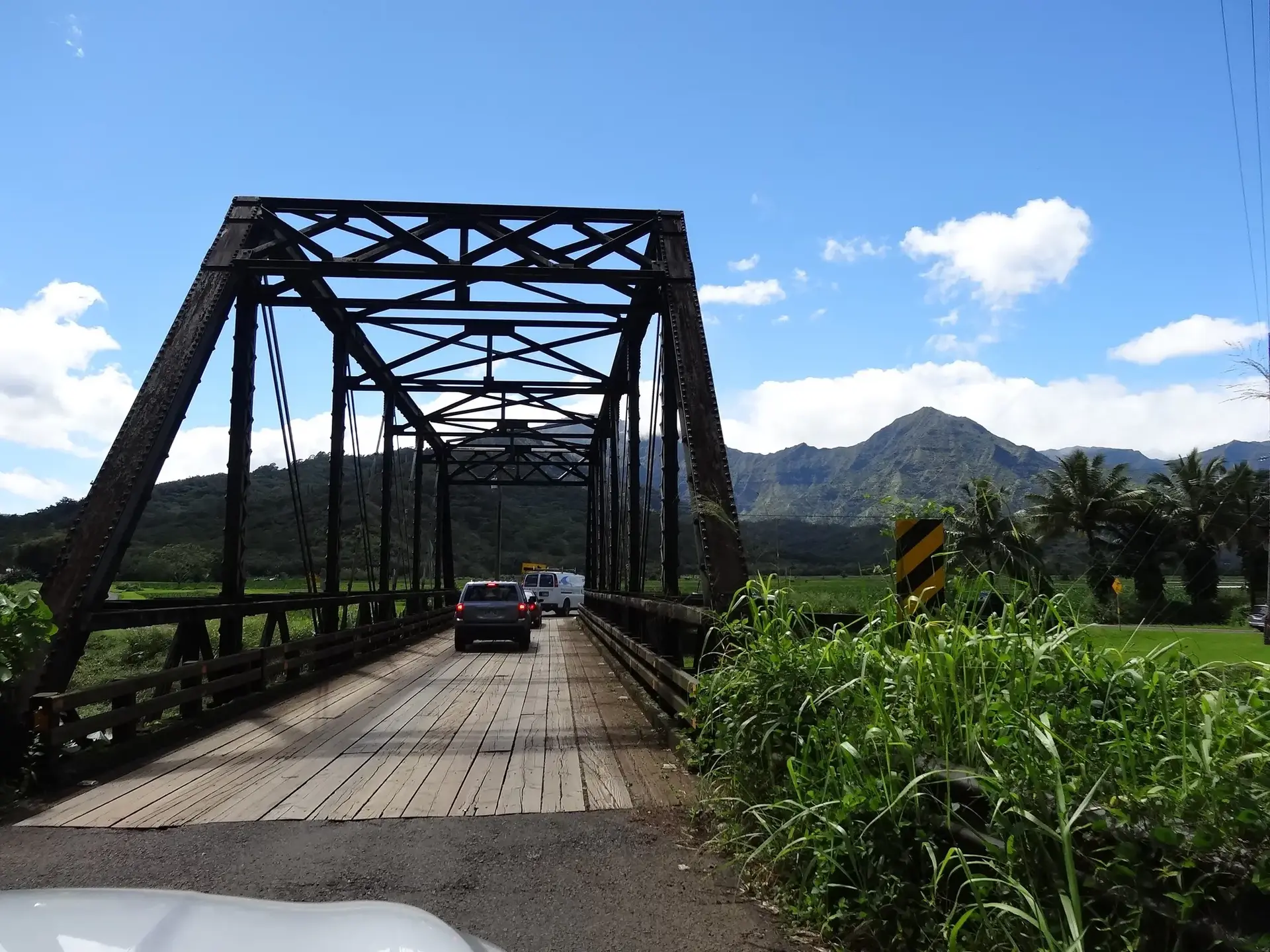 Cars cross an old metal bridge with green mountains in the background.
