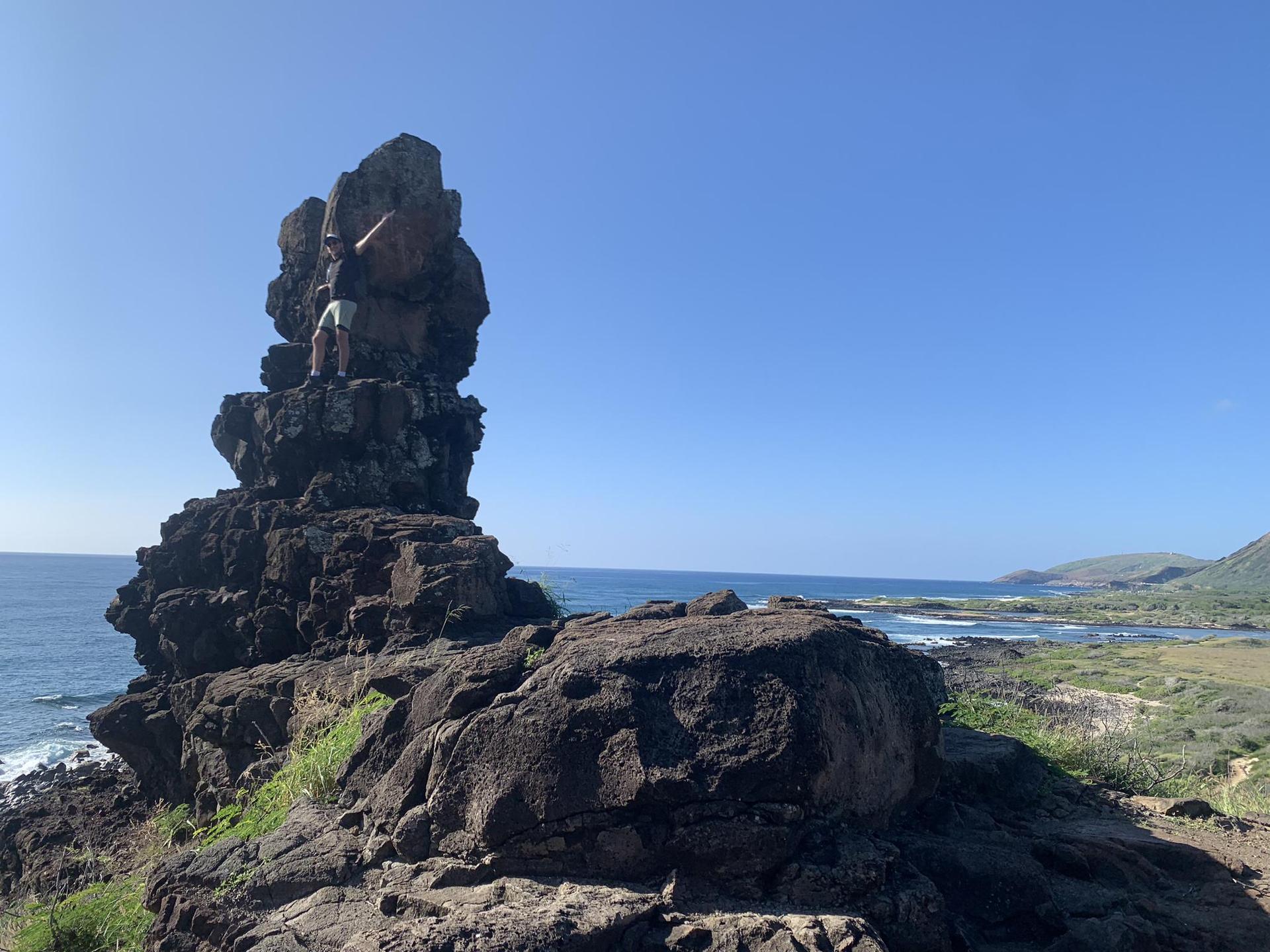 Person climbing a tall dark rock formation by the bright blue ocean.