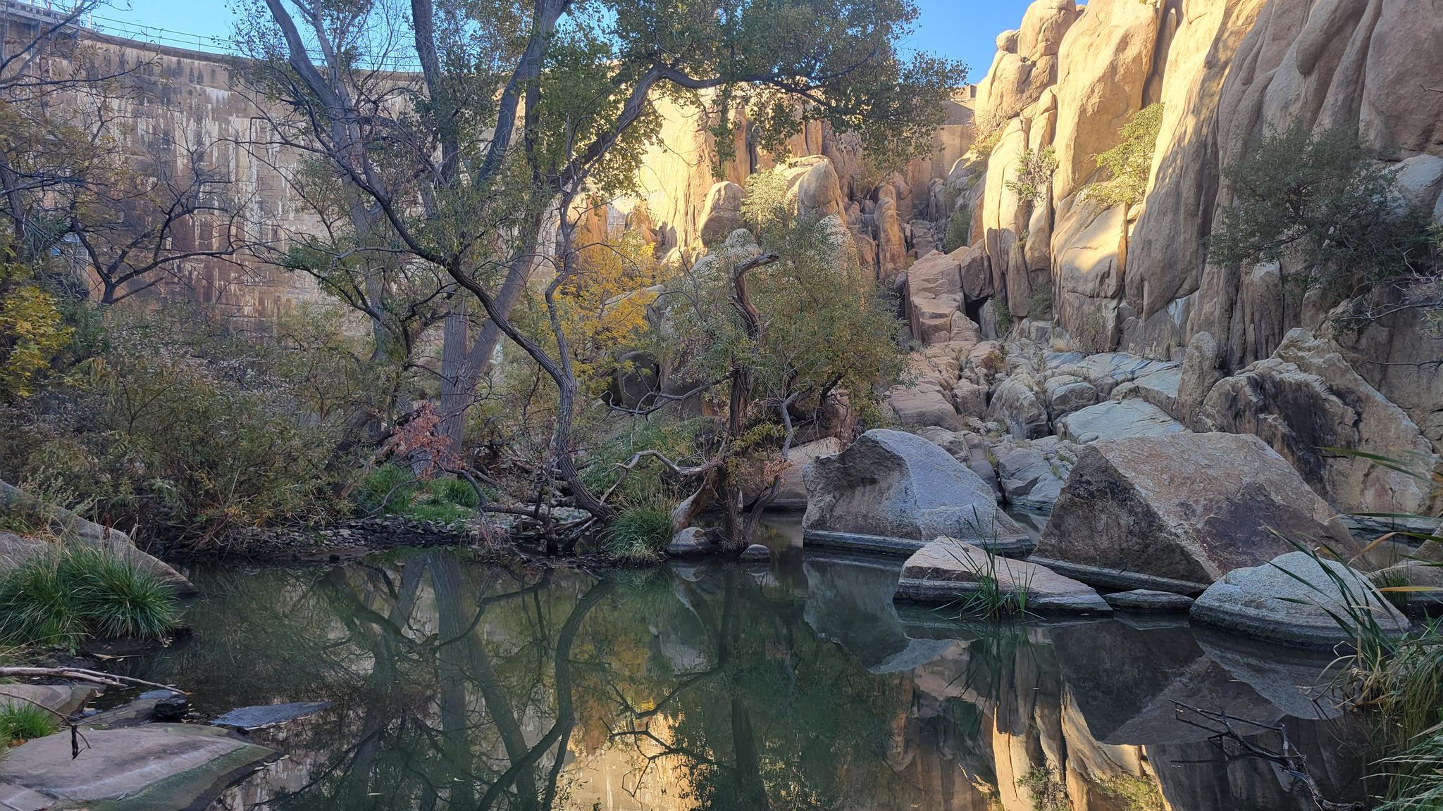 Calm water reflecting trees and sunlit rock formations, with a dam.