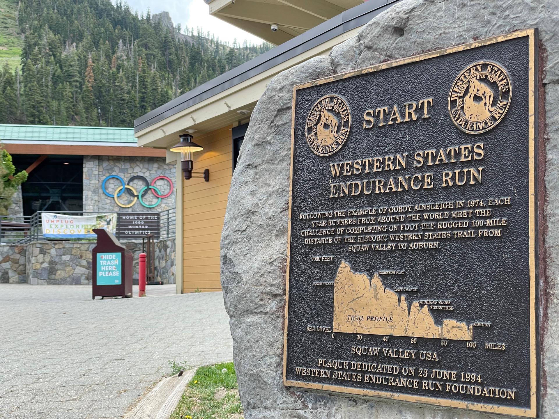 Bronze plaque on a rock next to a building, with Olympic rings visible.