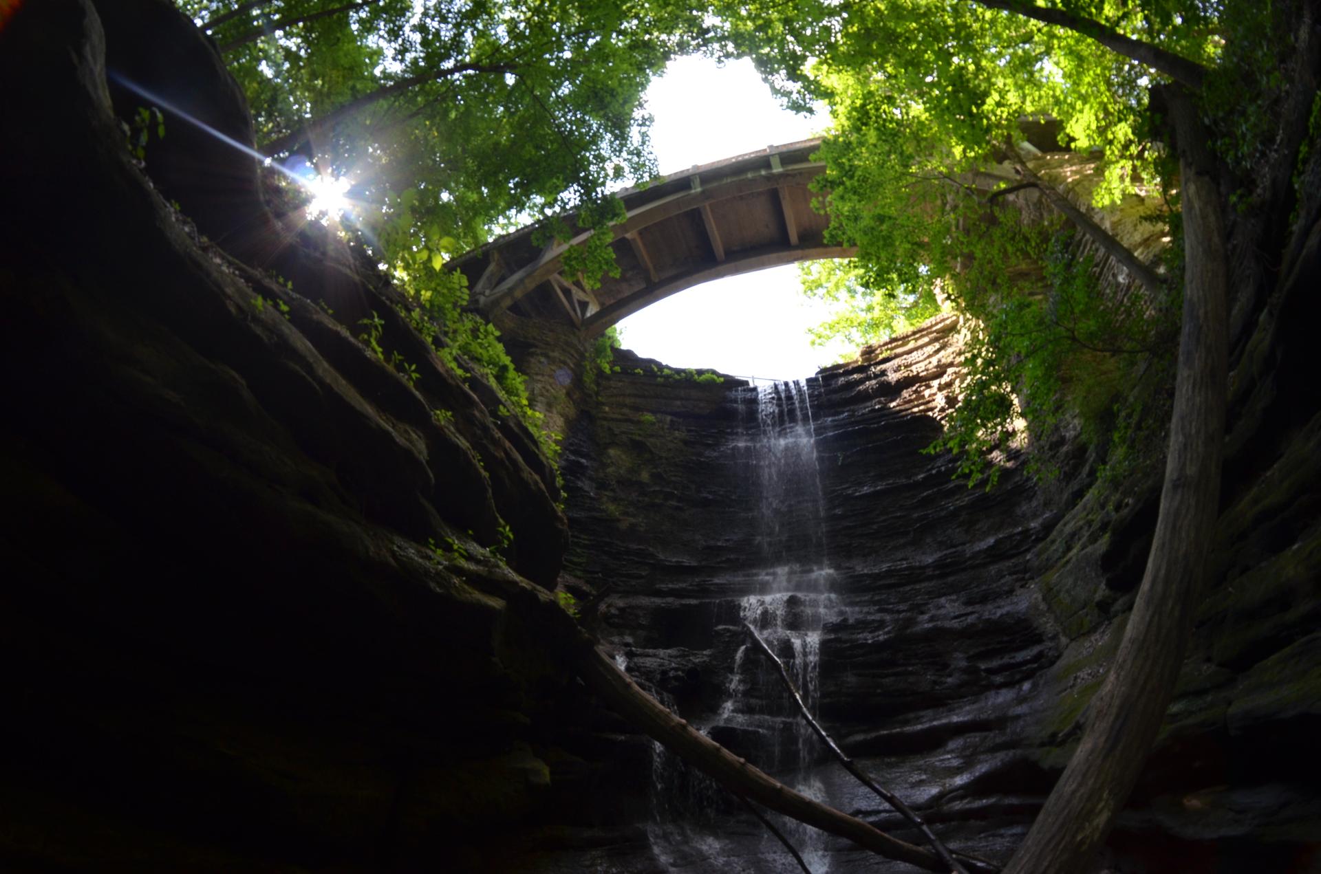 Waterfall cascades from an arched bridge opening, sun flares through green trees.
