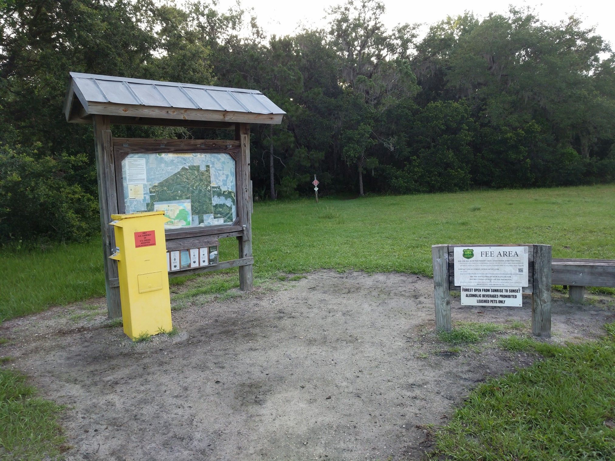 A wooden information board and yellow payment box stand by a grassy field. - Kolokee Loop Trail