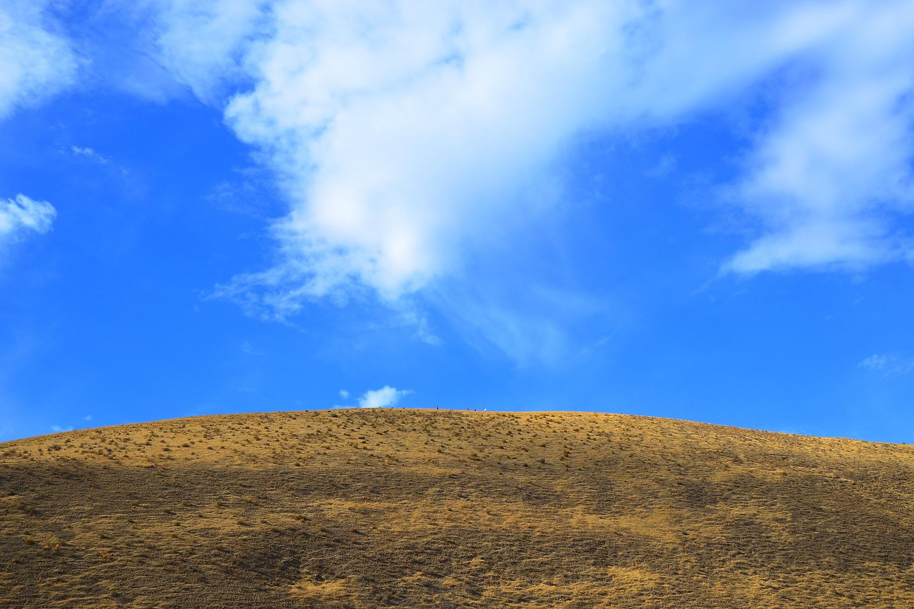 Bright blue sky with white clouds over a sunlit, brown grassy hill.
