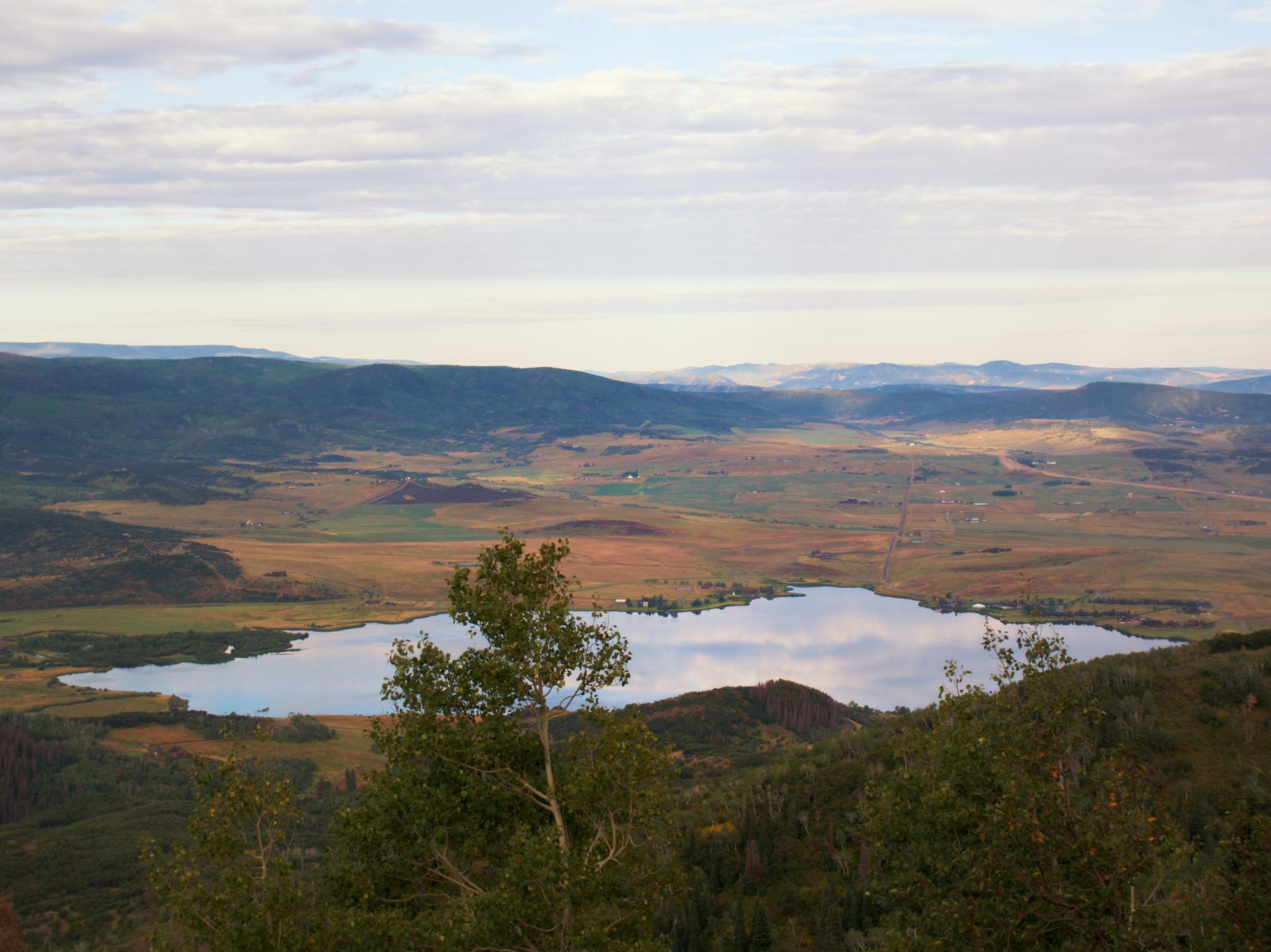 Expansive view of a calm lake reflecting cloudy sky, surrounded by mountains.