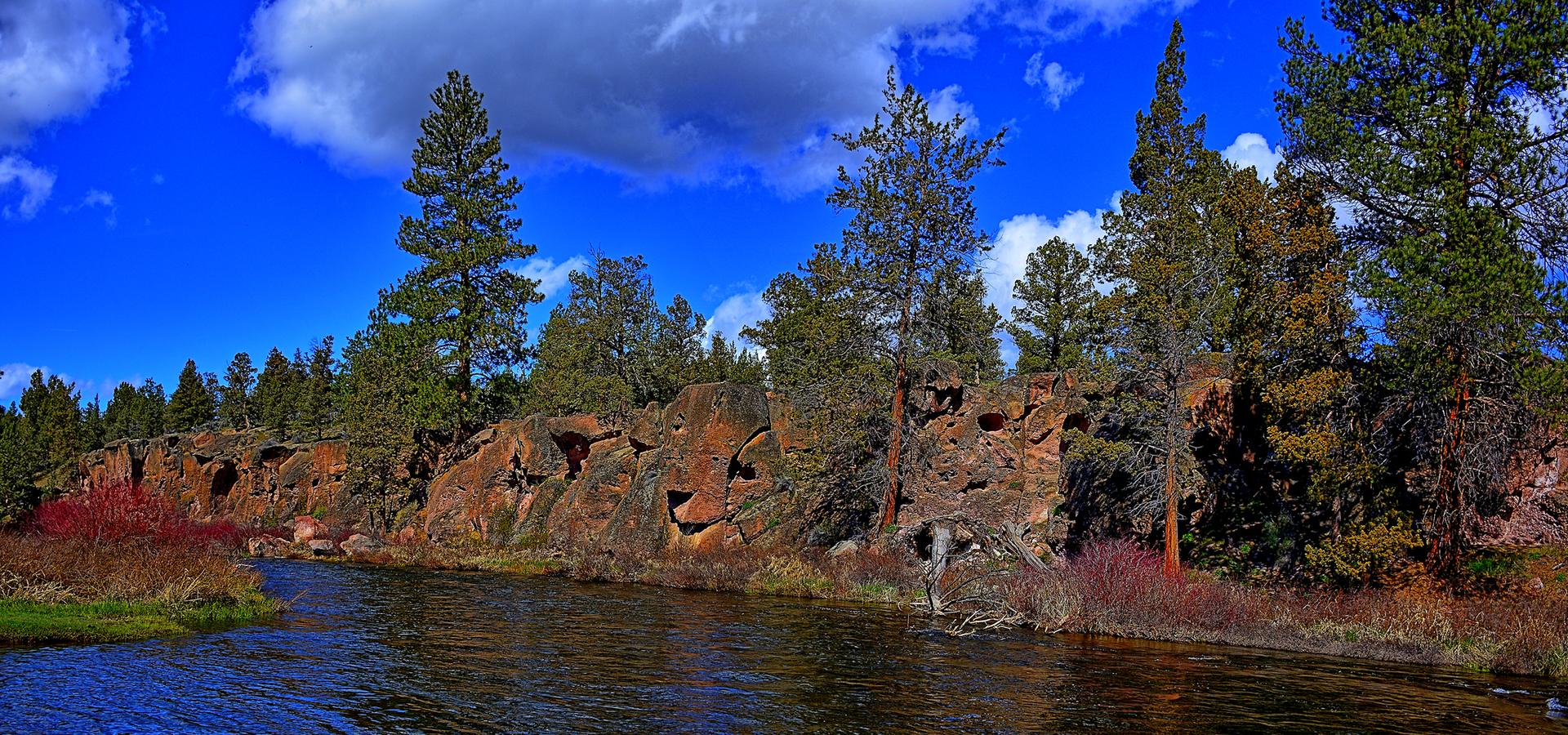A river flows past rocky cliffs and tall pine trees under a cloudy sky.